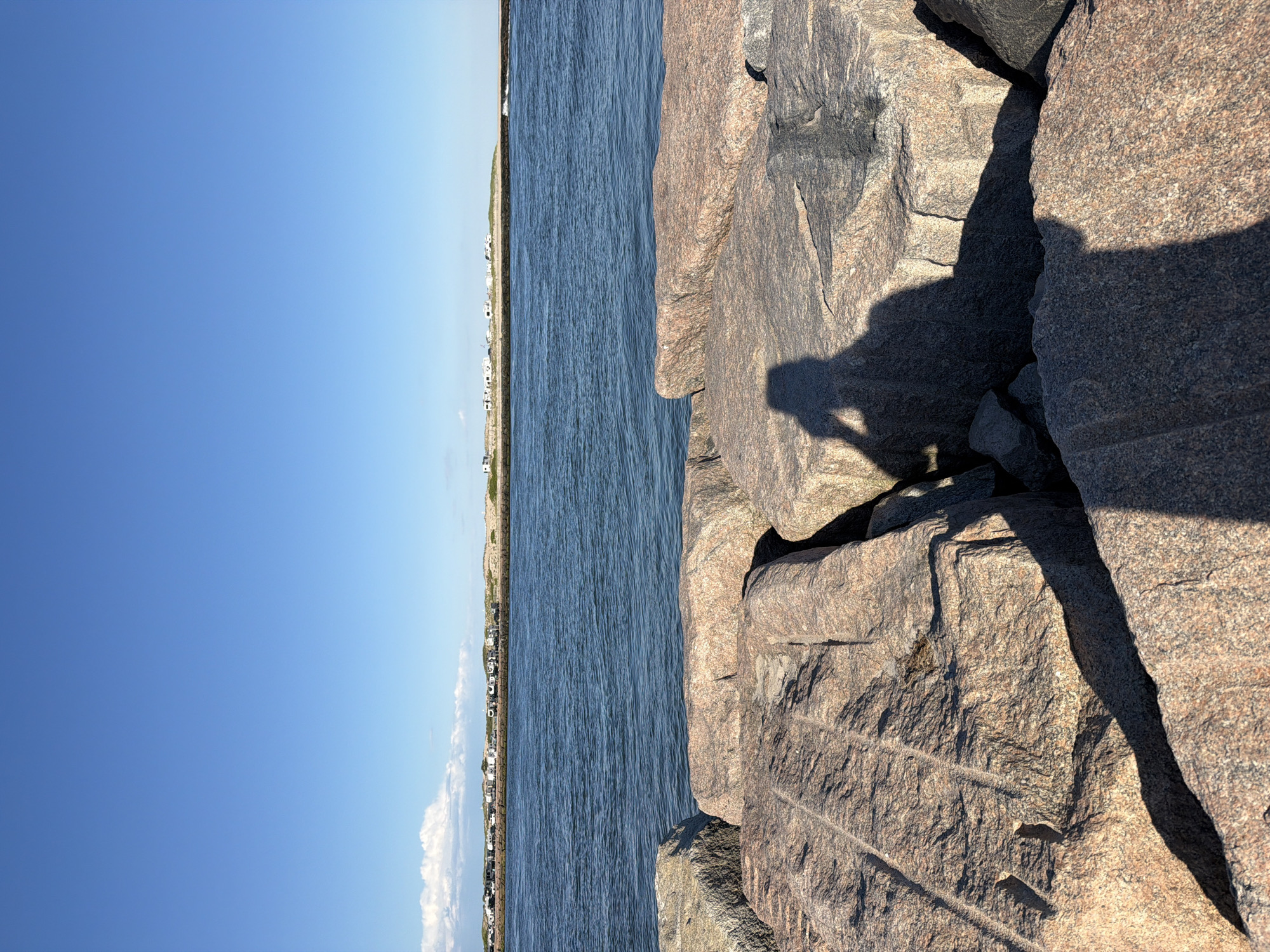 A shadow of a person holding a phone to take a picture is cast on large, rough-hewn granite rocks in the foreground. The rocks are of various sizes and shapes, some appearing to be part of a jetty or breakwater. Beyond the rocks, a body of calm, blue water stretches towards a distant shore. On the far shore, a line of sand dunes is visible, topped with vegetation and a row of recreational vehicles or campers. A clear, bright blue sky with a few wispy white clouds dominates the upper portion of the image. The scene appears to be set in a coastal area, likely Hampton Bays, United States of America, given the presence of a jetty, ocean, and beachside camping.