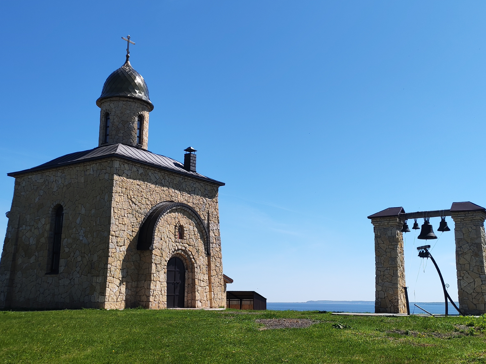 The image shows a stone chapel with a dome topped with a cross, located in Икское Устье, Russia. Next to the chapel, there is a structure with hanging bells. The chapel and bell structure are situated on a grassy area overlooking a body of water. The sky is clear and blue. There are no people visible in the image. A small shed is visible in the background.