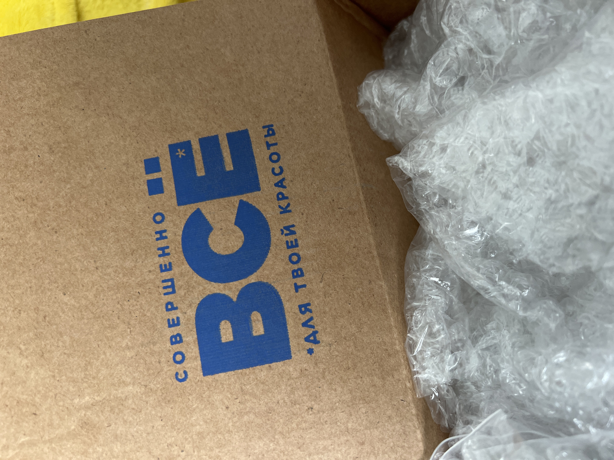 The image shows a cardboard box filled with bubble wrap. The side of the box displays a logo and text in blue. The text reads "СОВЕРШЕННО" (SOVERSHENNO), then "BCE" (ВСЕ), and finally "*ДЛЯ ТВОЕЙ КРАСОТЫ" (*DLYA TVOEY KRASOTY) (FOR YOUR BEAUTY). Considering the text is in Russian, the image is likely taken in Russia. "ВСЕ" (VSE) chain store exists in Chaikovsky, Russia. The background of the shot is somewhat out of focus, with what appears to be a yellow object in the upper left.