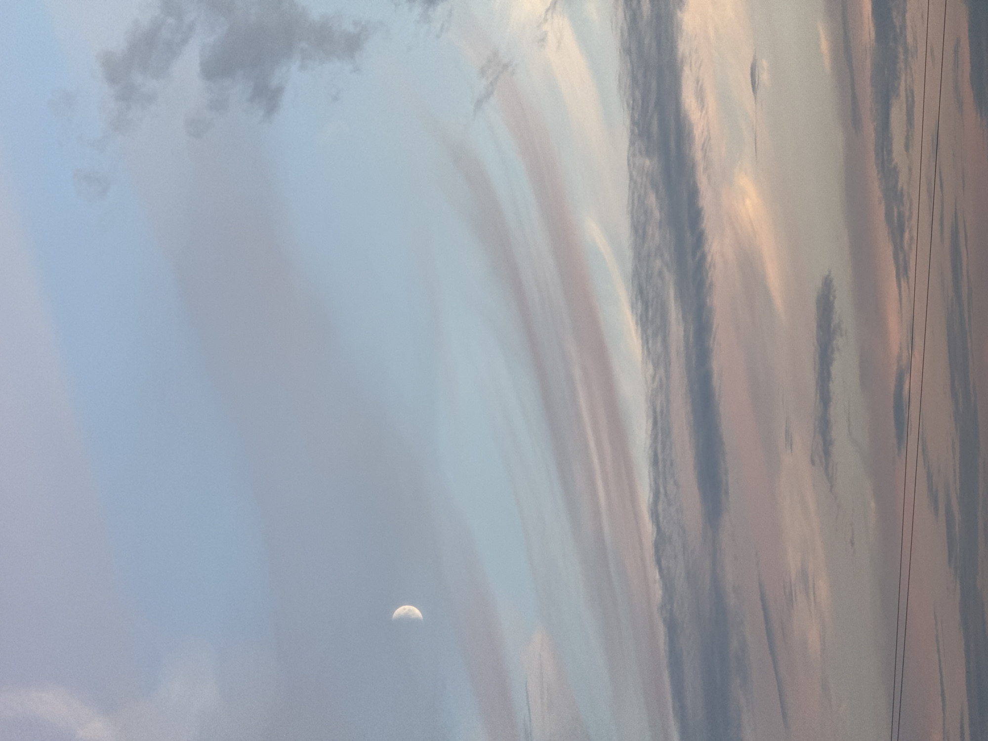 The image captures a twilight sky with wispy clouds painted in soft hues of pink, blue, and gray. A crescent moon is visible in the upper left quadrant. In the lower portion of the image, two thin, dark horizontal lines, likely power lines, are present, stretching across the frame. The background consists of a gradient of colors, transitioning from a pale blue at the top to softer pinks and grays towards the bottom. The scene appears to be an outdoor, natural environment, possibly overlooking a landscape or city at dusk. No people or specific actions are depicted. The location is identified as Sovkhoza imeni Lenina, Russia, suggesting a rural or semi-rural setting.