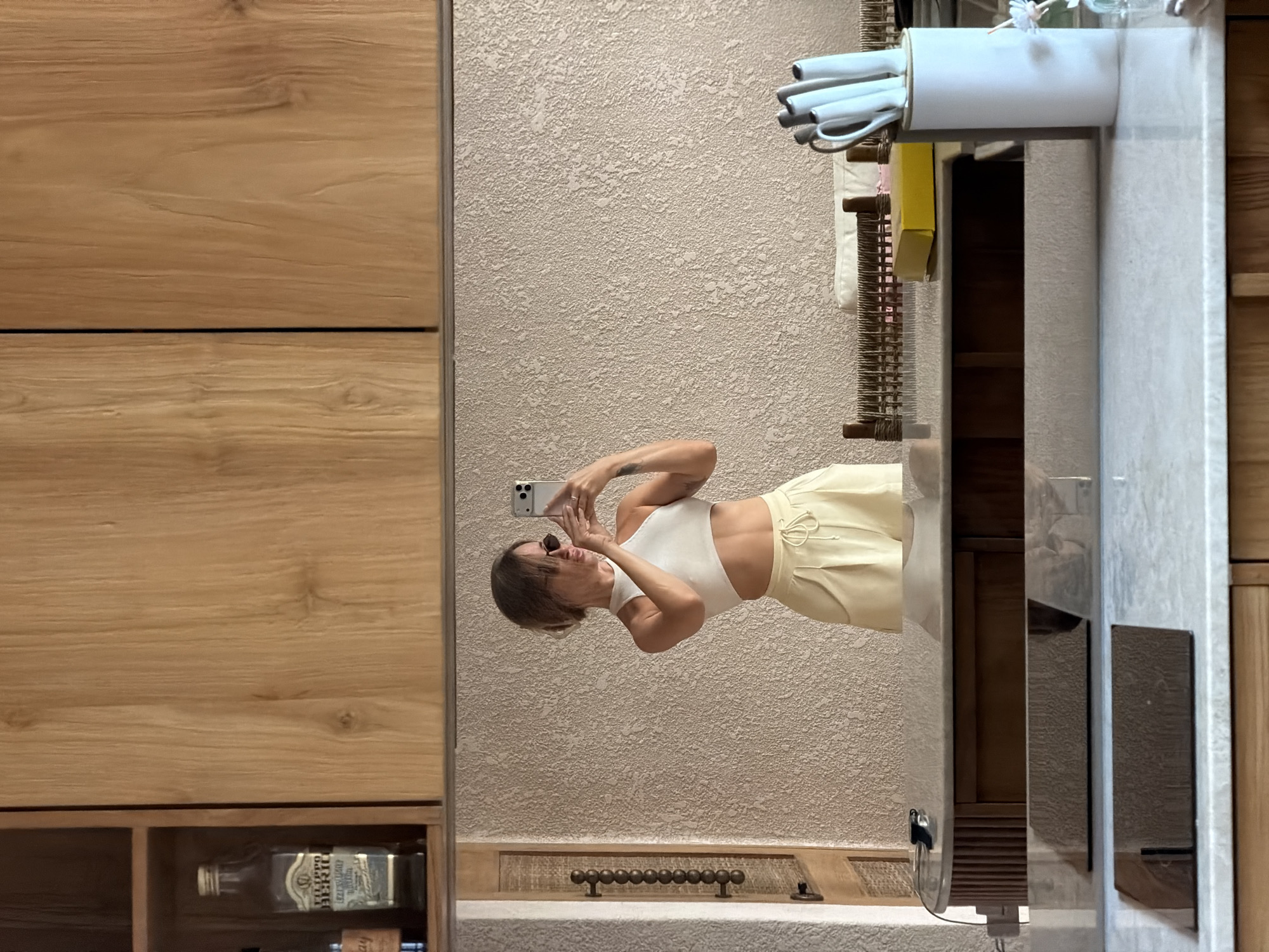 The image is a vertically oriented, slightly overexposed photograph taken in a bathroom with light beige textured walls and light wood cabinetry. In the foreground, a person is reflected in a large mirror. The person, a woman with brown hair and sunglasses, is wearing a white crop top and light yellow wide-leg pants. She is holding a smartphone and appears to be taking a selfie, with her arms raised and one hand touching her face. Her expression is playful and she seems to be smiling.
The bathroom features a modern aesthetic. To the left of the mirror, light wood cabinets are visible, with one shelf containing a glass bottle of Filippo Berio olive oil. Above the mirror, a white holder with several light blue items, possibly hair styling tools, is mounted on the wall. The mirror itself reflects a shelf containing wooden slats. Below the mirror, a textured countertop is visible with a decorative metallic element that resembles a string of beads.
The lighting suggests it could be daytime, possibly indoors with artificial light contributing to the overall brightness. The overall mood conveyed is casual and relaxed. No specific time of day or weather conditions are discernible. The location context is specified as Mengwi, Indonesia.