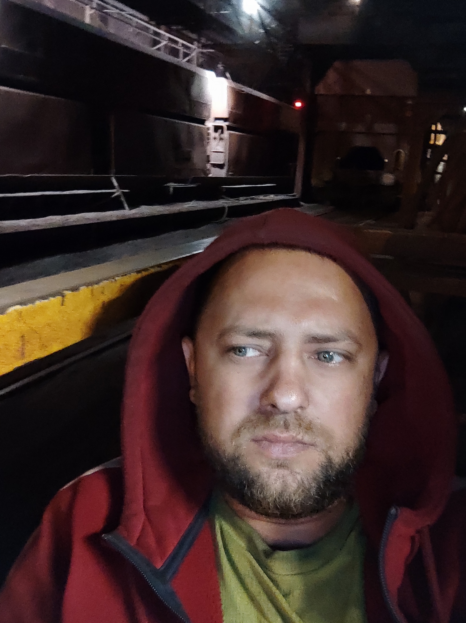 A close-up, dimly lit selfie captures a man from the chest up, wearing a red hooded sweatshirt and a green t-shirt. He has light blue eyes, a light complexion, and a short beard. The background appears to be an industrial setting, possibly a steel mill in Salzgitter, Germany. Structures and beams are visible, some illuminated by artificial light, creating a stark and somewhat ominous atmosphere. A yellow stripe is visible on a lower surface in the foreground.