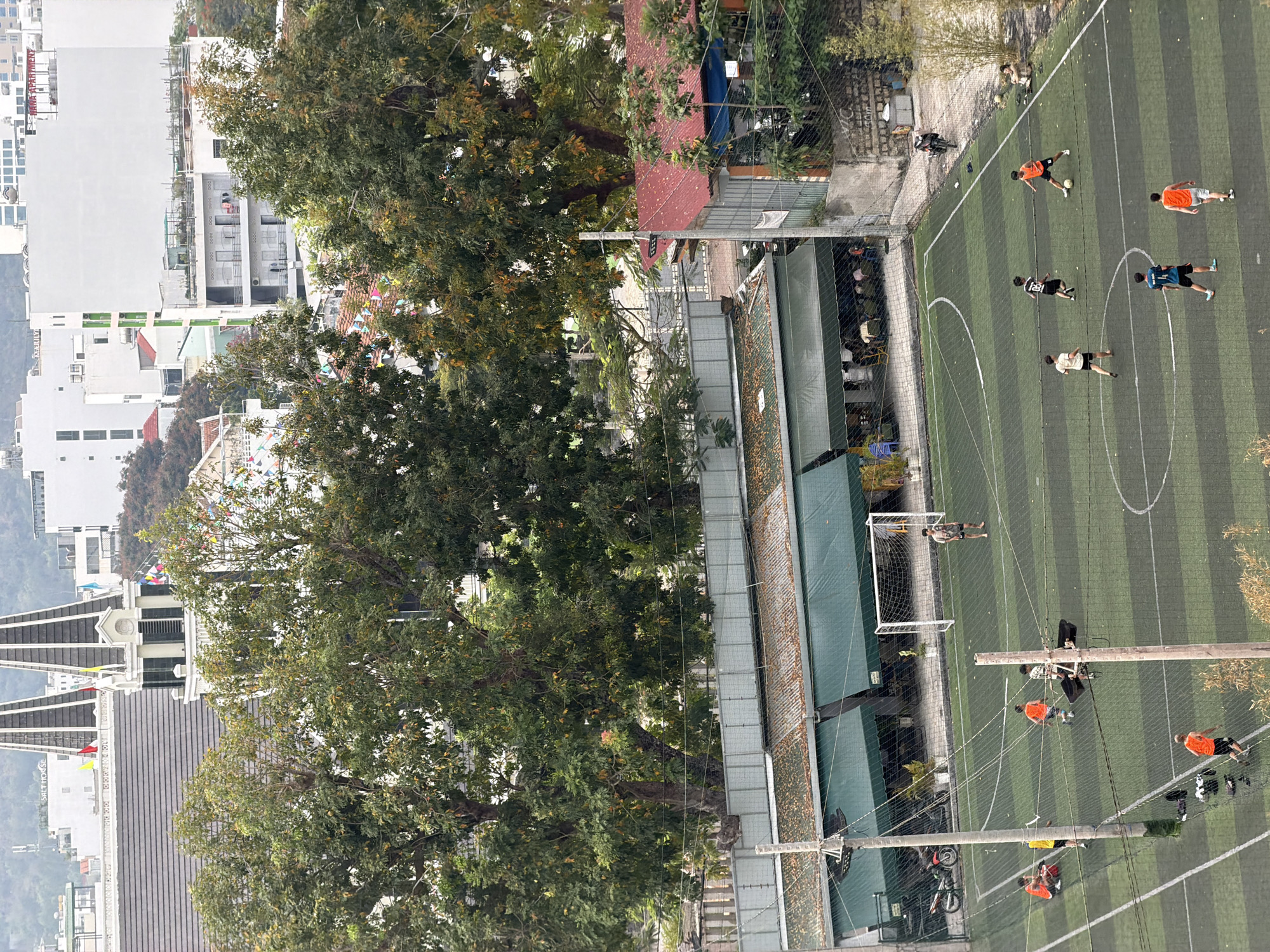 An elevated, rotated view shows a portion of Bac Nha Trang Ward, Vietnam. Foregrounded is a tree with dense foliage obscuring some of the urban landscape. Behind it is a partially visible soccer field with multiple individuals, some appearing to be playing soccer while others are observing. A section of buildings are situated to the left of the tree. Power lines and poles traverse the soccer field. Various building types are visible, including high-rise structures.