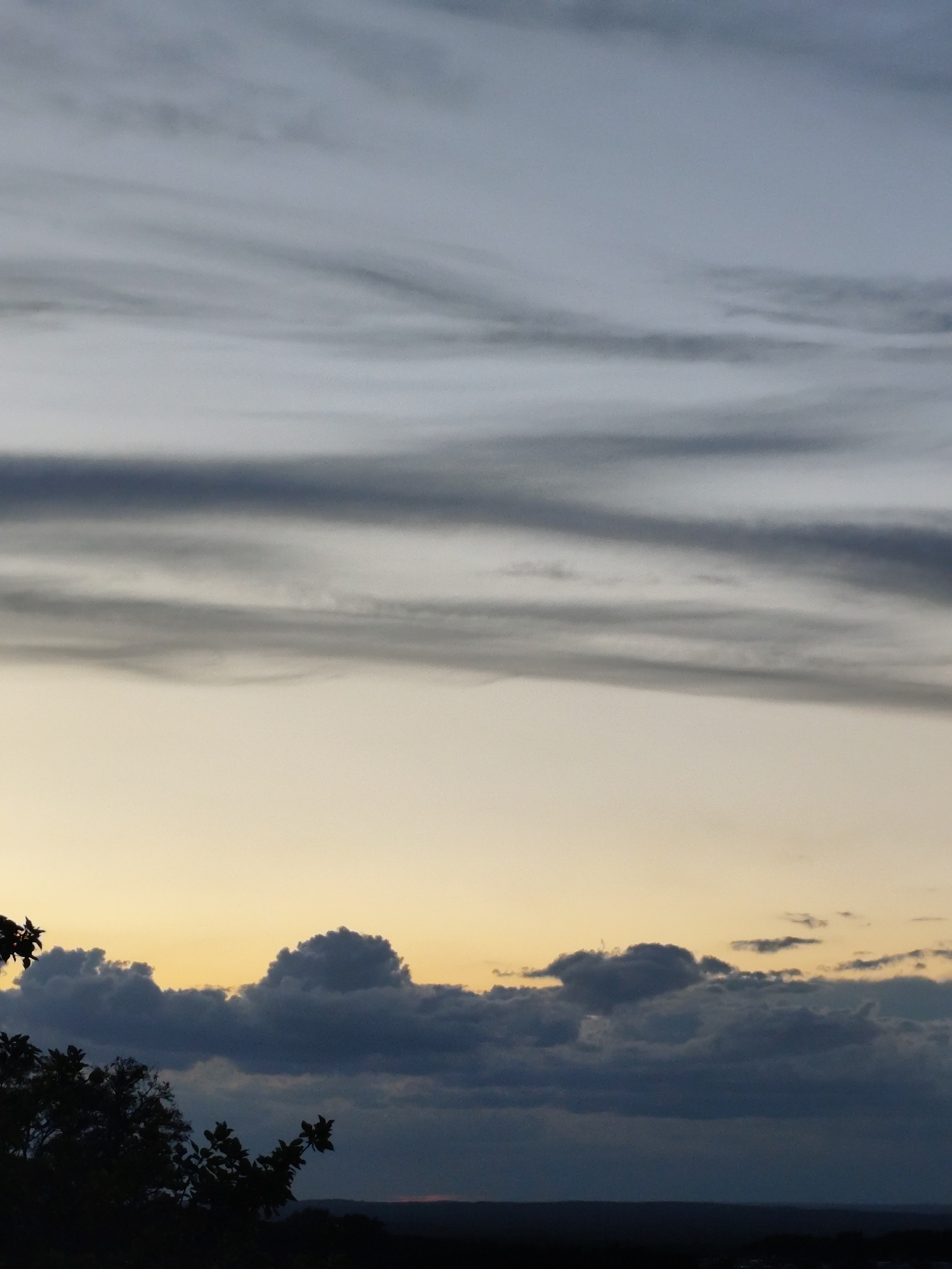 A sky photograph shows a gradient of light from the horizon to the top of the image. The horizon is dark, with silhouettes of trees and possibly buildings. Above this, there is a band of soft yellow and orange light, suggesting either sunrise or sunset. Higher up, large, dark grey clouds are clustered, with some lighter grey clouds above them. The top of the image is a pale blue-grey sky with horizontal, wispy grey clouds. The image appears to be taken from an elevated position, overlooking a landscape. The specific location is not identifiable from the image content.