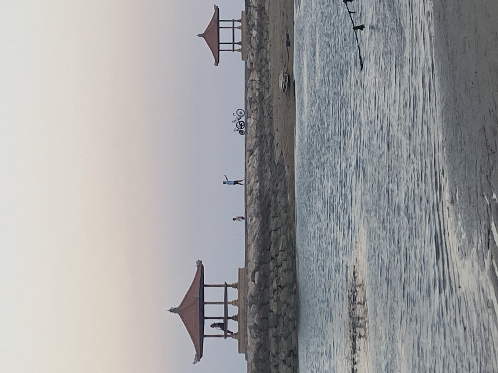 The image depicts a serene coastal scene at what appears to be sunset or sunrise in Denpasar, Indonesia. In the foreground, calm water ripples gently against a sandy shore. Extending from the shore into the water is a man-made structure, a stone jetty or causeway, topped with a pathway.
On the jetty, two distinct Balinese-style gazebos are visible. The first gazebo on the left has a tiered, reddish-brown roof and is supported by pillars. Inside, a solitary figure is seated, appearing relaxed. Further along the jetty, near the second gazebo, a person in a blue shirt is captured in mid-gesture, possibly waving or pointing. Two bicycles are parked beside this individual. The second gazebo, on the right, is similar in architectural style to the first.
The background is a soft, hazy sky transitioning through pale pinks and blues, indicative of twilight. The overall atmosphere is peaceful and tranquil. The presence of the gazebos and the architectural style strongly suggest a location within Denpasar, possibly a beach or a harbor area popular for recreational activities.