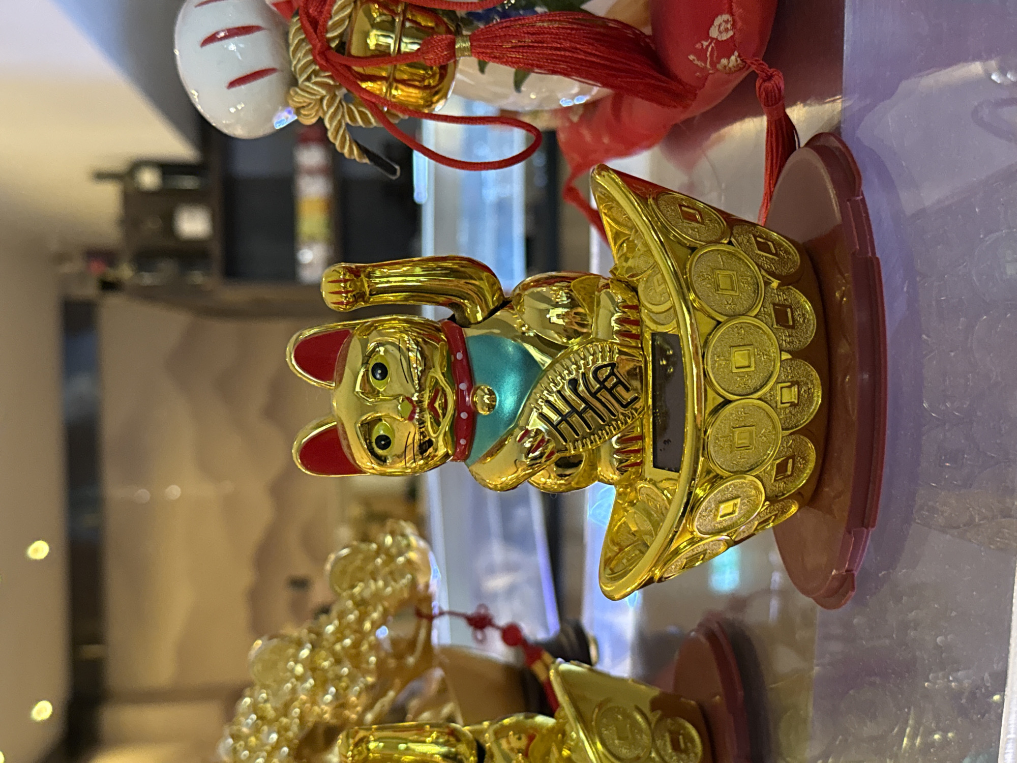 A close-up, eye-level shot shows a gold-colored lucky cat figurine with its left paw raised in a welcoming gesture. The cat is seated in a gold-colored boat filled with gold coins. The figurine is positioned on a dark red circular base. In the background, out of focus, are more gold decorations, shelves with bottles, and warm-toned wall art. The scene appears to be indoors, possibly in a shop or restaurant. The image was taken in Nordhausen, Germany.