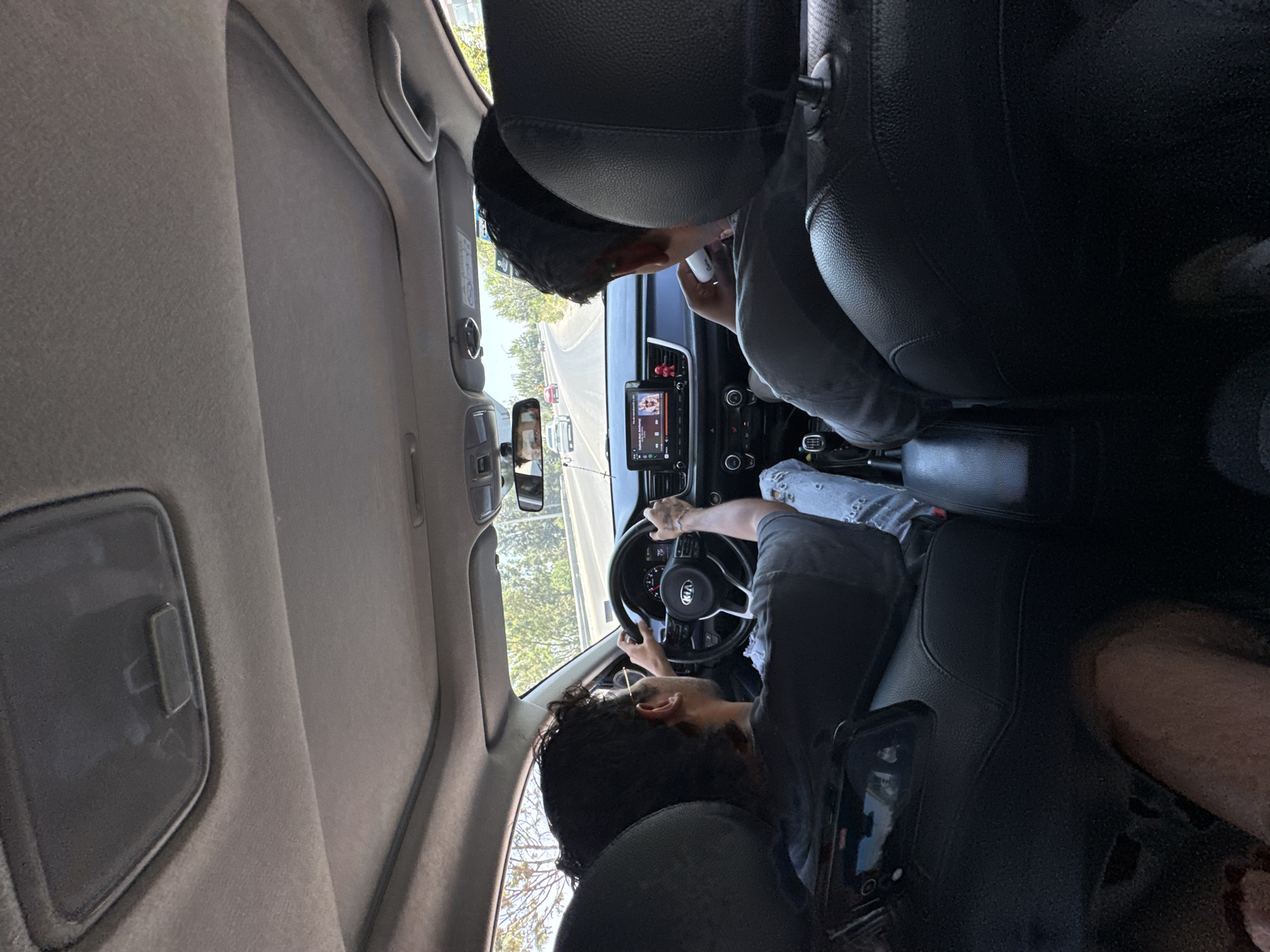 The image captures the interior of a car from the perspective of a backseat passenger, looking forward. Two individuals are visible in the front seats. The person in the driver's seat, with dark curly hair and a beard, is wearing a dark t-shirt and has both hands on the steering wheel, which features a "KIA" logo. Their left forearm displays tattoos.
To their right, a passenger, wearing a grey long-sleeved shirt and a baseball cap, is seen holding a white object, possibly a phone or cup, and appears to be looking down at it. A cell phone displaying what looks like a social media interface is resting on the side of the passenger seat.
The car's dashboard is visible, featuring a central infotainment screen. The screen shows the Spotify application, displaying a song titled "Canción para las bestias" by "La Renga." A small red, possibly bobblehead, figure is attached to the dashboard above the screen.
Through the windshield, a clear, sunny daytime road is visible, curving slightly to the left, with trees and other vehicles in the distance. The rearview mirror reflects parts of the car's interior. The car's headliner is light-colored, and sun visors and grab handles are discernible.
The overall scene depicts a casual drive during the day in San Bernardino Tlaxcalancingo, Mexico.