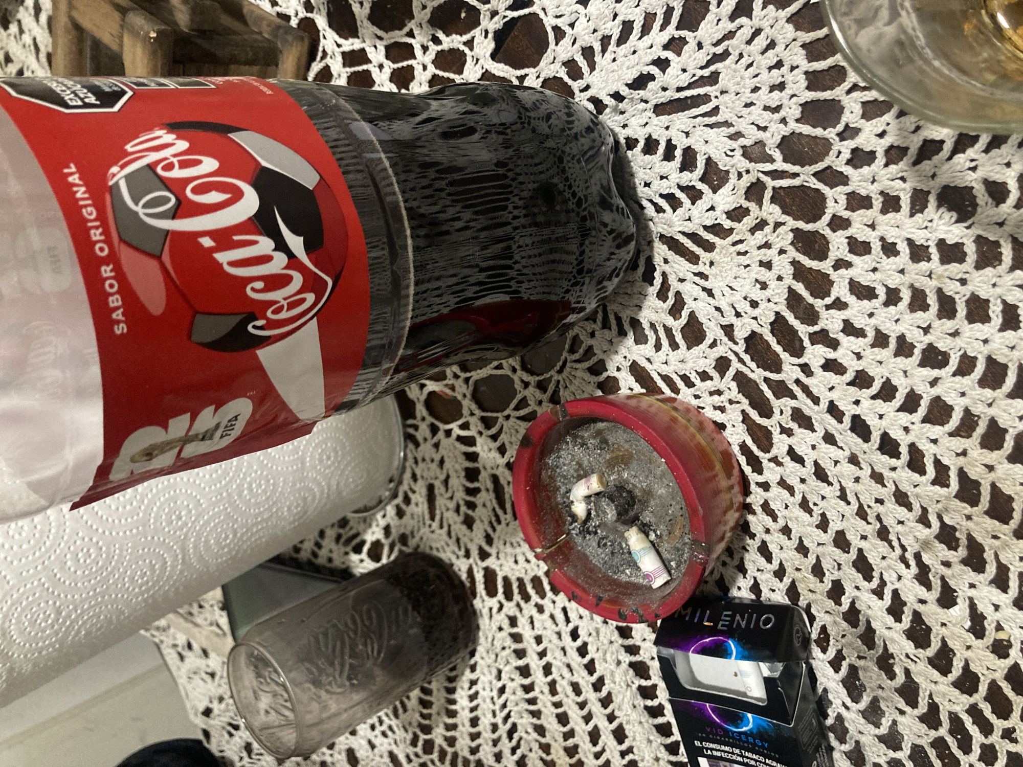 The image shows a still life scene arranged on a white crocheted doily. The items include a large plastic bottle of Coca-Cola with a red and white label, a roll of paper towels, a used glass Coca-Cola bottle, an ashtray filled with cigarette butts and ash, and a black cigarette pack labeled "MILENIO VID ICERGY." The scene appears to be indoors on a dark brown surface. The presence of "FIFA" on the bottle of coca-cola suggests this item may be related to a sporting event. Given the language used on the Coca-Cola and cigarette packaging, the objects suggest a location where Spanish is spoken. The brand "MILENIO" is associated with Argentina. It is impossible to know if this is in Buenos Aires, Argentina, however, it could be.