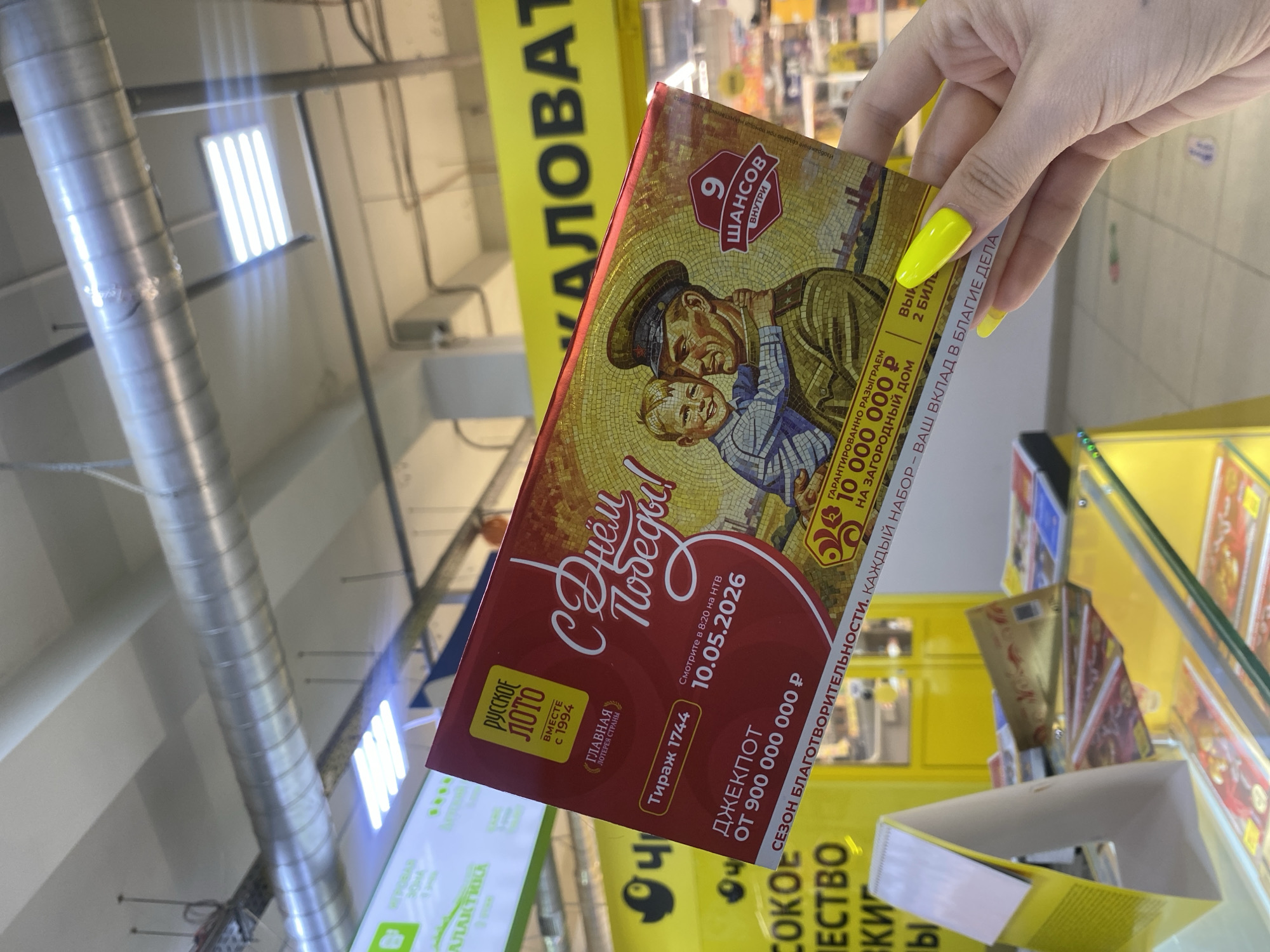 Here is a description of the image:
The image is taken indoors, seemingly in a store in the city of Chaikovsky, Russia. A hand with long, pointed nails painted in bright yellow is holding a red-and-gold Russian Lotto ticket celebrating Victory Day, with a drawing of a soldier embracing a child. The lottery offers 9 chances to win, with the drawing scheduled for May 10, 2026, and a jackpot of 900 million rubles. The lottery guarantees 10 million rubles will be given out. In the background, other similar lottery ticket packages are visible. The environment suggests a store aisle with product displays. Overhead, industrial-style lighting fixtures and pipes are visible. A yellow sign with black Cyrillic text is partly visible on the left side of the frame. The foreground is occupied by the lottery ticket.