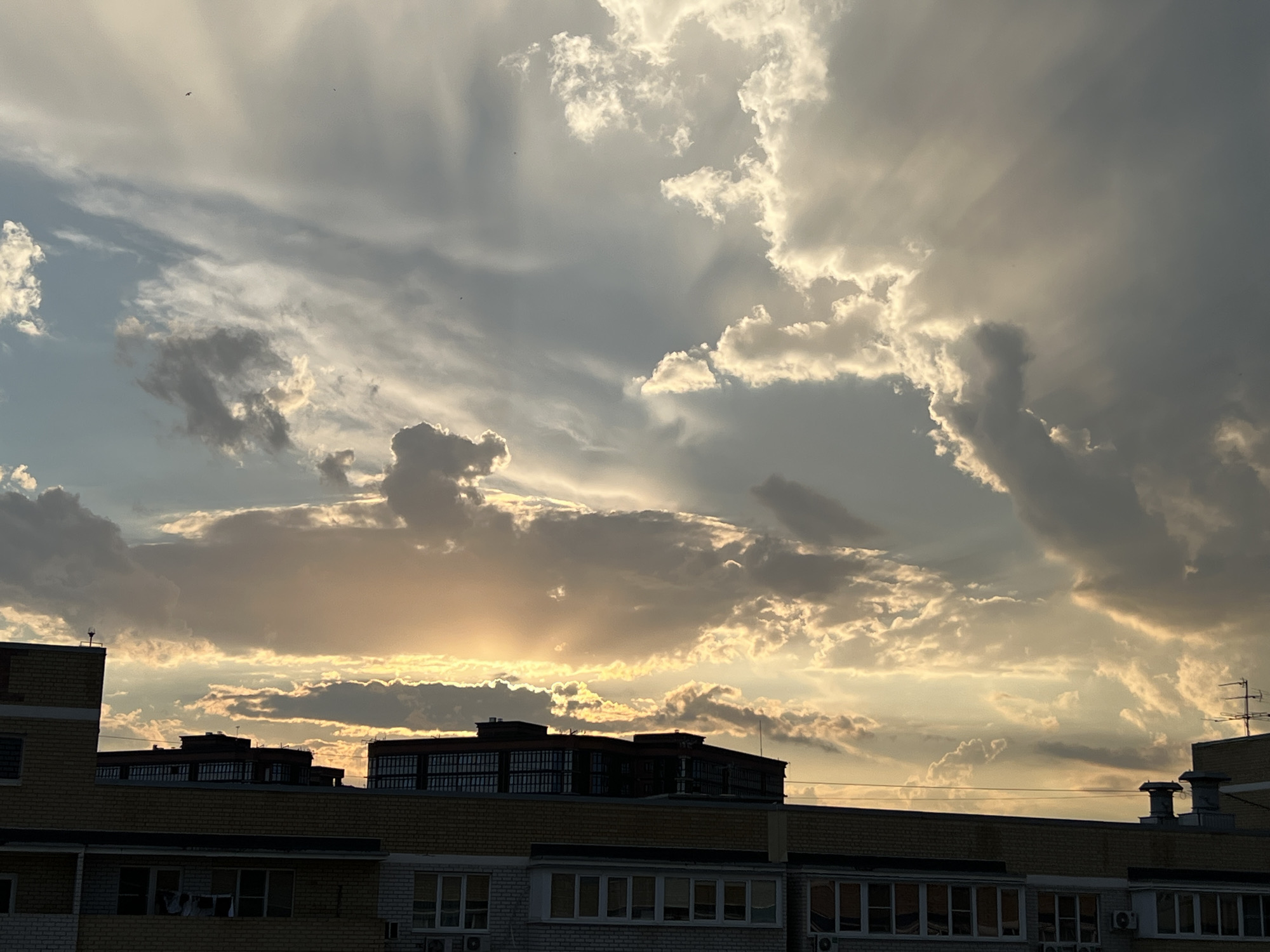 This media file captures a dramatic sunset over a cityscape, likely in Krasnodar, Russia. The scene is dominated by a vast sky filled with dynamic clouds, illuminated by the setting sun. Shafts of light pierce through the darker cloud formations, creating a sense of depth and highlighting the texture of the clouds. The sun itself is partially obscured by a band of clouds, casting a warm, golden glow across the sky.
In the foreground, the silhouettes of apartment buildings are visible. These structures, with their brick facades and rows of windows, suggest an urban environment. Some buildings have visible antennas and ventilation units, indicating residential use. The architecture suggests a typical residential area in a Russian city.
The time of day is clearly sunset, with the low angle of the sun and the warm, diffused light. The weather appears to be partly cloudy, with both dark storm clouds and bright, sunlit clouds present. The interplay of light and shadow creates a somewhat dramatic and awe-inspiring atmosphere. The vastness of the sky and the grandeur of the cloud formations evoke a sense of wonder and peace. There is no visible text in the image. The overall impression is one of a beautiful, natural spectacle occurring within a mundane urban setting.