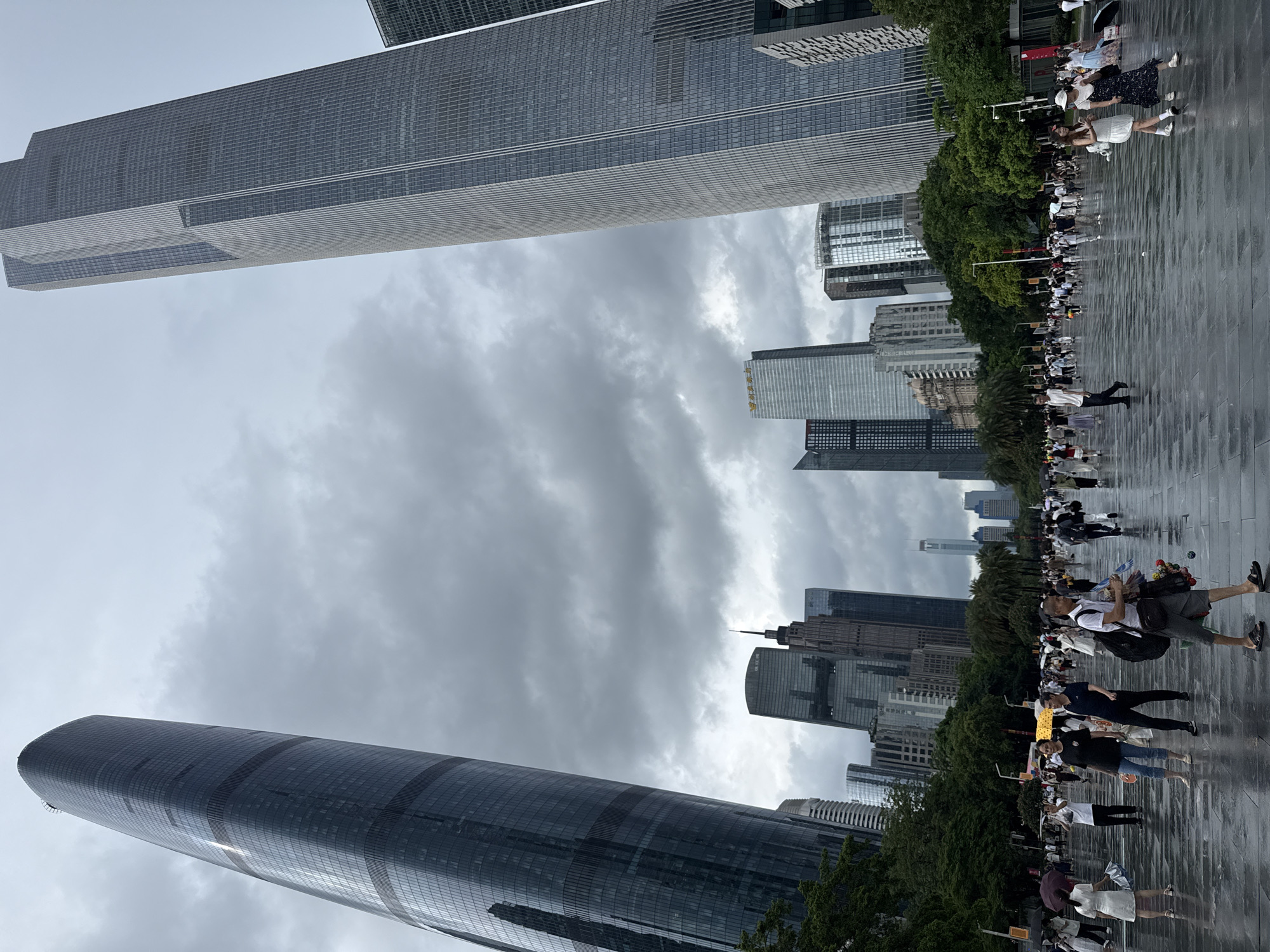 This image depicts a wide, wet plaza in Tianhe District, Guangzhou, China, with a crowd of people walking. The plaza's surface is reflective, indicating recent rain. Towering skyscrapers dominate the background, with two particularly prominent glass-faced buildings on the left and right sides of the frame, angled towards the viewer. The sky is overcast with dark, dramatic clouds. The crowd is diverse, with individuals and small groups spread across the plaza. Some people are holding umbrellas, suggesting continued light rain or a recent shower. Several individuals are carrying bags or belongings. Trees and other greenery are visible behind the plaza, interspersed with more urban structures and smaller buildings. The scene appears to be a public space in a modern, bustling city center.