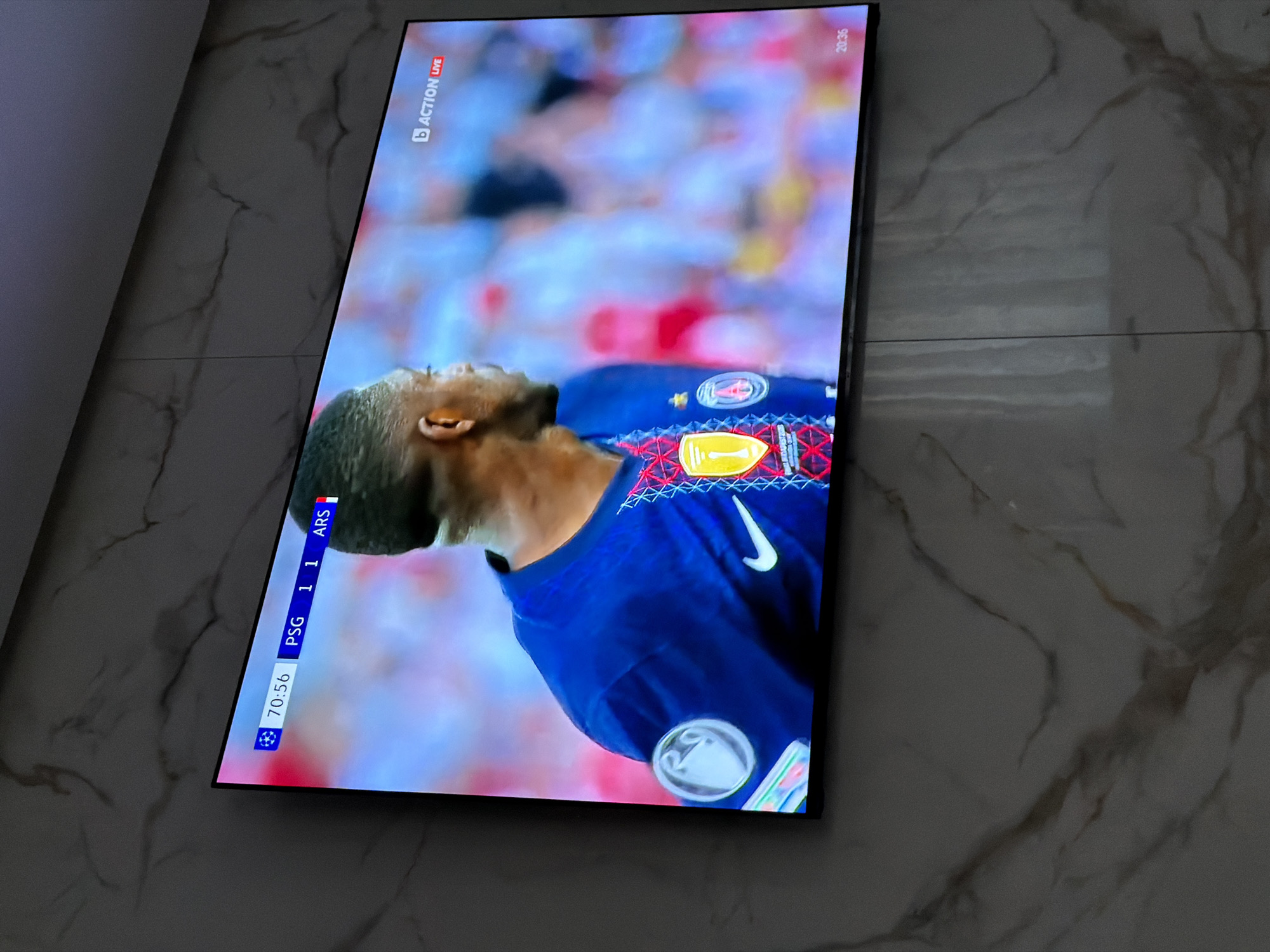 A television screen shows a football game between PSG and Arsenal, with a score of 1-1 at 70:56. A player in a blue jersey is prominently displayed on the screen. The Action Live logo is visible in the top left corner, and "20:35" is displayed in the top right. The TV is mounted on a light-colored wall with a marble-like pattern. The image was taken indoors. The city of Asenovgrad, Bulgaria is not discernible from the visual information provided.