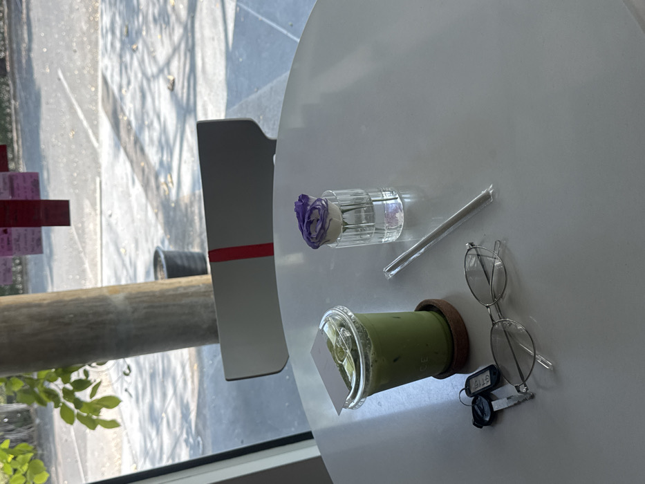 A white table displays a green beverage with a plastic lid and a white card inserted, along with a straw, a glass cylinder holding a purple flower, eyeglasses, and keys. The table is positioned near a window. A gray chair with a red stripe is visible. Outside the window, green foliage and a glimpse of a building or structure can be observed.