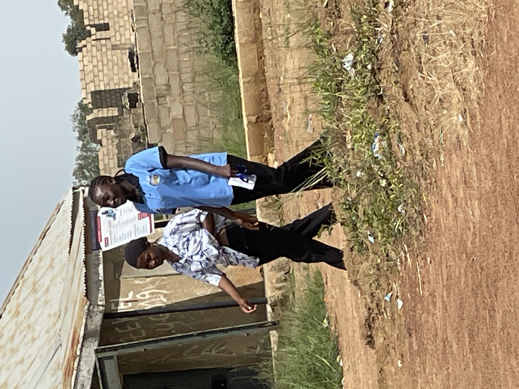 The media file depicts two young men outdoors, possibly in Durumi, Nigeria. The man in the foreground is wearing a blue shirt and black pants, and appears to be holding a document or a small book. He is looking down and to his left. Behind him and slightly to his right, another young man is visible from the waist down, wearing a patterned shirt and black pants. He is also looking downwards.
The setting appears to be a somewhat unkempt area. To the left of the men is a building with a corrugated metal roof and a brick wall. There's a poster or notice board on the wall of this building, with visible text in a foreign language. The ground is a mix of dirt and patchy green grass, with some scattered debris. In the background, more brick structures are visible, along with trees. The lighting suggests it's daytime, with no strong shadows, indicating possibly overcast weather or mid-day sun. The overall atmosphere seems casual, possibly related to education or some form of community gathering given the presence of the poster.