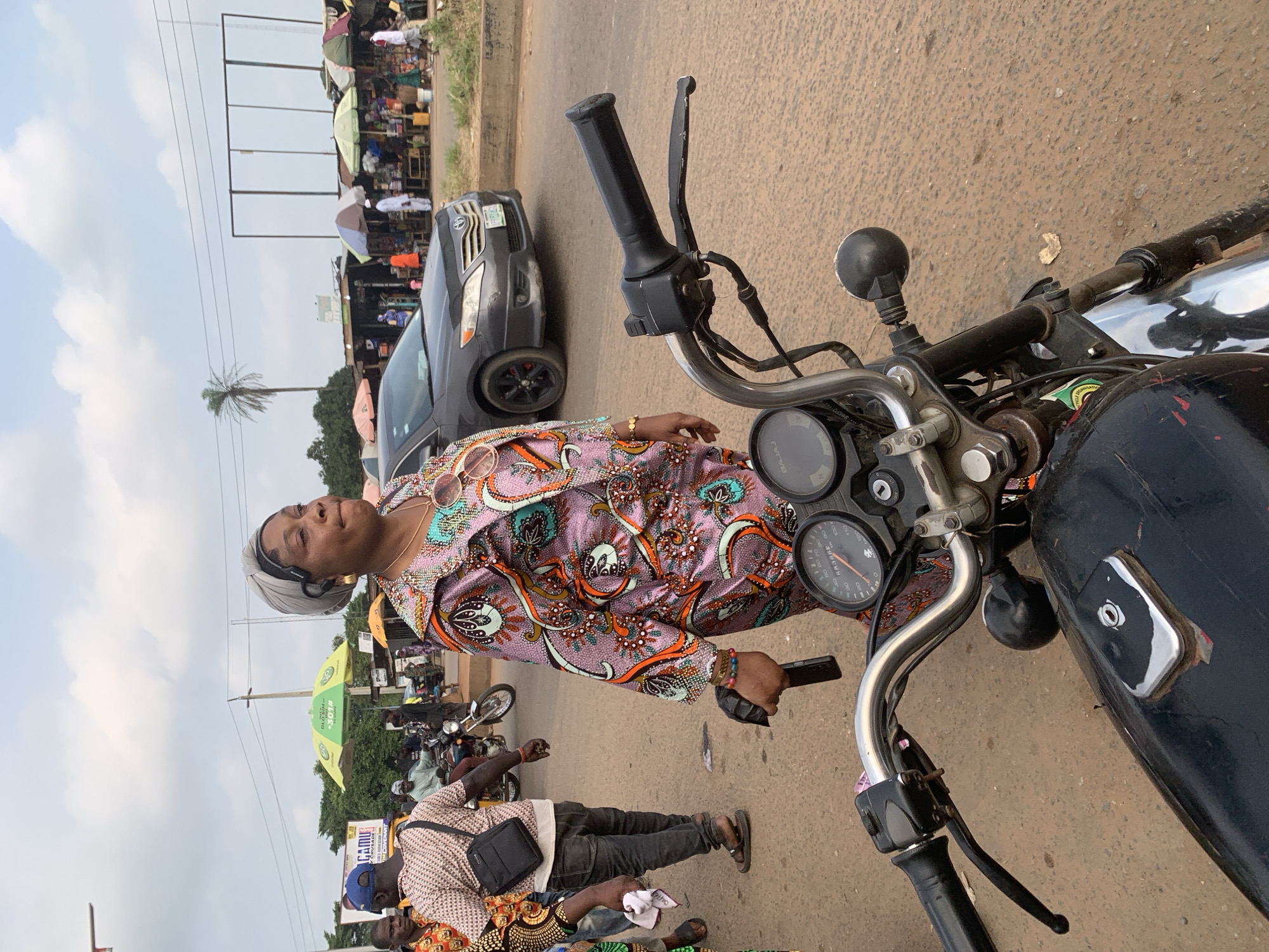 A medium shot shows a woman in the foreground, standing next to a motorcycle on a dusty road. She is wearing a patterned purple and pink dress with an orange and green design, a grey headwrap, and large gold hoop earrings. She is holding a dark object in her right hand. The motorcycle handlebars are visible in the bottom foreground. In the mid-ground, other people are present, some on motorcycles, and a grey car is parked on the side of the road. The background features market stalls with various wares displayed, some covered by colorful umbrellas. A palm tree and utility poles are visible against a cloudy sky. The scene appears to be a bustling street market in Shagamu, Nigeria.