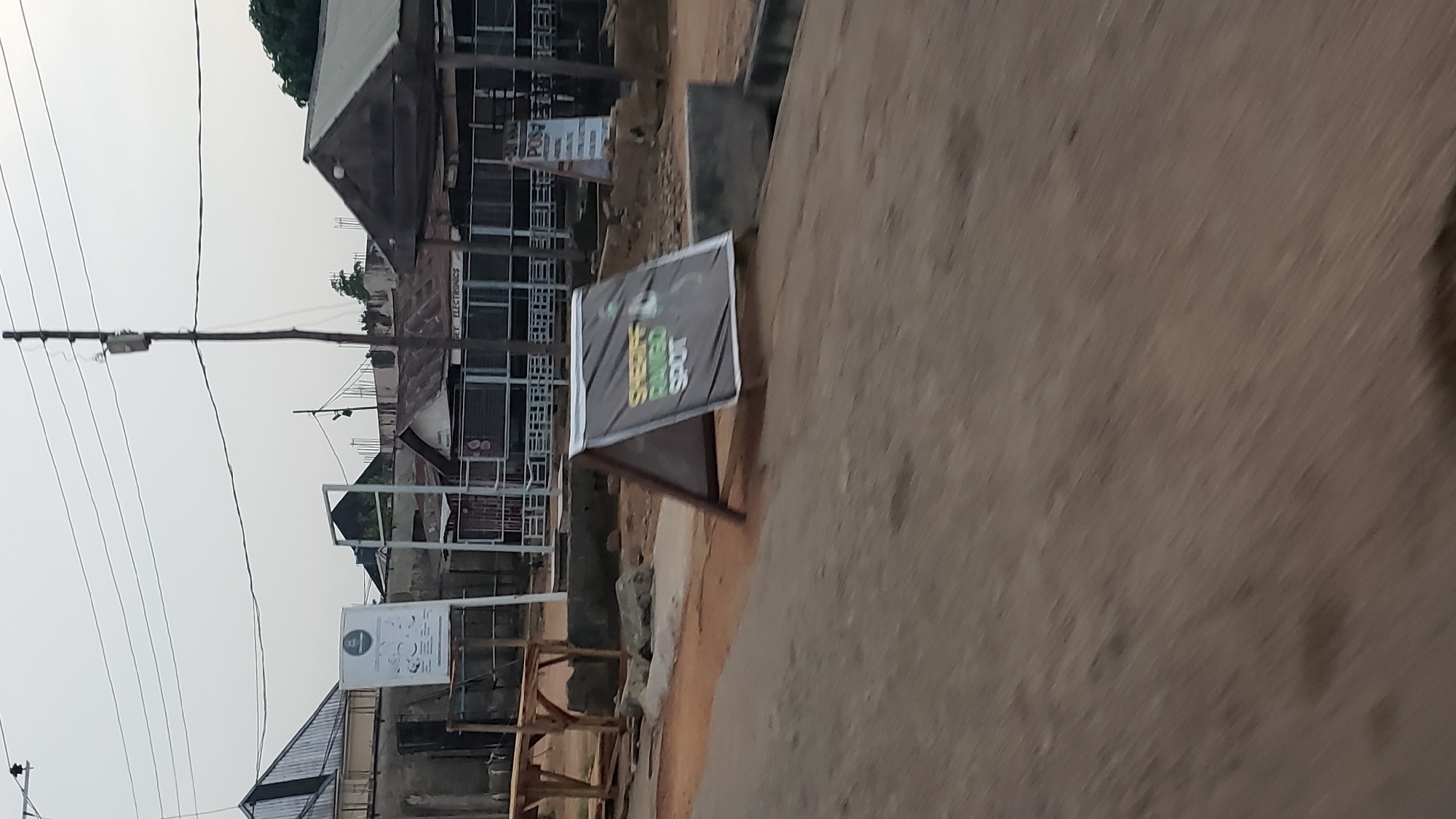 The image depicts a street scene in Jalingo, Nigeria, likely in an urban or semi-urban area. The foreground is dominated by a rough, dusty road surface. To the left, a wooden structure, possibly a market stall or seating area, is visible. In the middle ground, an A-frame sign stands prominently, displaying text and a graphic that appears to advertise "SHERIFF BINGO SPOT." The sign is dark-colored with lighter text and green graphics.
Behind the sign, several buildings with corrugated iron roofs are visible, suggesting a commercial or residential area. One building has signage that reads "POS" and another appears to be a shop with the words "BET ELECTRONICS" partially visible. Utility poles and power lines crisscross the sky, indicating the presence of electricity infrastructure. The overall environment appears somewhat weathered and rustic. There are no people clearly visible in the image. The sky is overcast, suggesting a cloudy day.