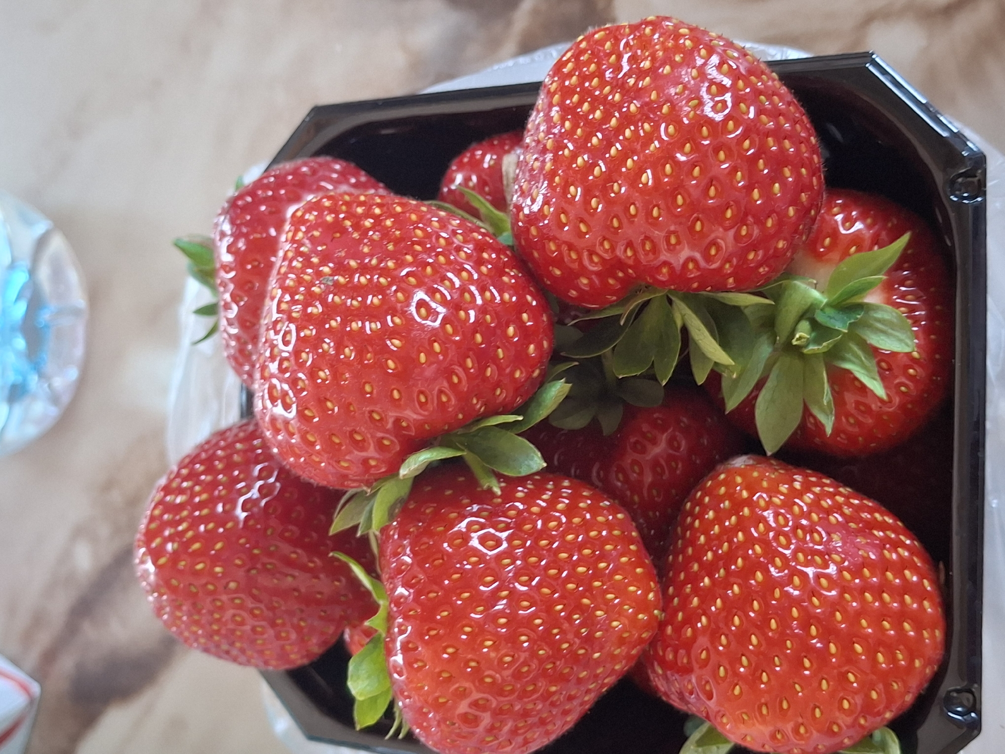 A black plastic clamshell container is filled with fresh, bright red strawberries. The strawberries are plump and juicy, with visible seeds and green stems. They are arranged in a cluster, with some overlapping. The background is a blurry mix of a light-colored, textured surface and a hint of blue in the upper right corner, possibly a bottle or container. The lighting suggests an indoor setting. There is no discernible urban environment or specific location in Åsgårdstrand, Norway.