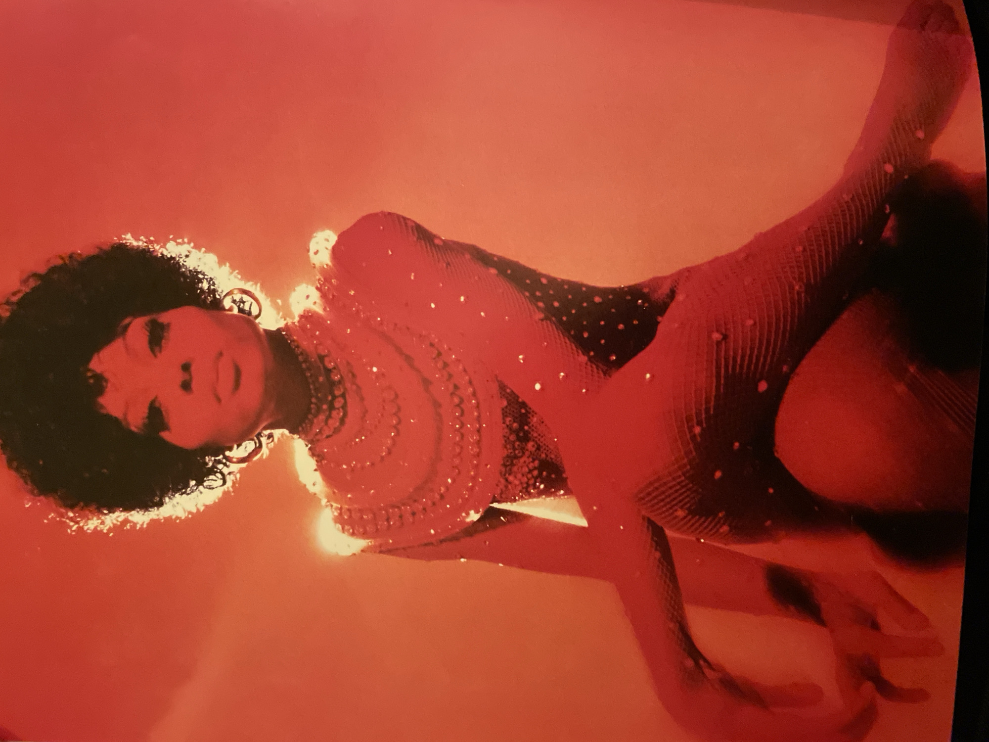 A studio portrait displays a person with dark, voluminous afro-textured hair, bathed in monochromatic red and orange lighting. The individual, presumed female, is adorned with large hoop earrings and a multi-layered necklace featuring prominent circular embellishments. They wear a form-fitting, sparkling bodysuit or jumpsuit, heavily encrusted with reflective rhinestones or sequins across the torso, arms, and legs. The garment on the limbs exhibits a textured, possibly fishnet, pattern. The subject is depicted in a dynamic, seated pose with bent limbs against a uniform red-orange background, characteristic of a controlled studio environment. This photograph was captured in Barcelona, Spain; however, no specific city landmarks or external environments are discernible within the image.