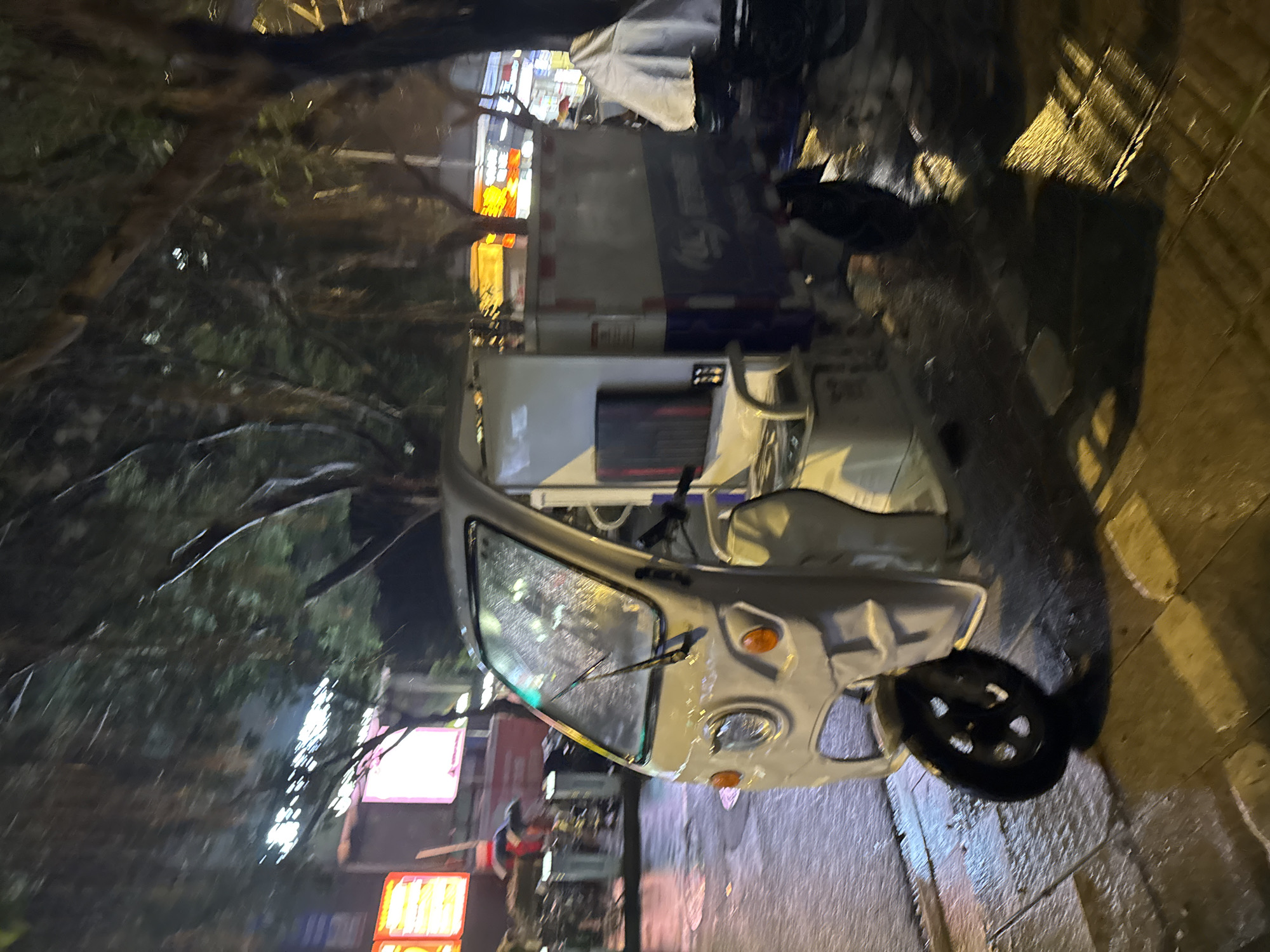 Observed in Tianhe District, China, is a three-wheeled vehicle parked on a cobblestone street at night. The vehicle is positioned near a sidewalk edge. Trees and illuminated commercial signs are visible in the background. A person is seen near the signs. The scene is dimly lit.