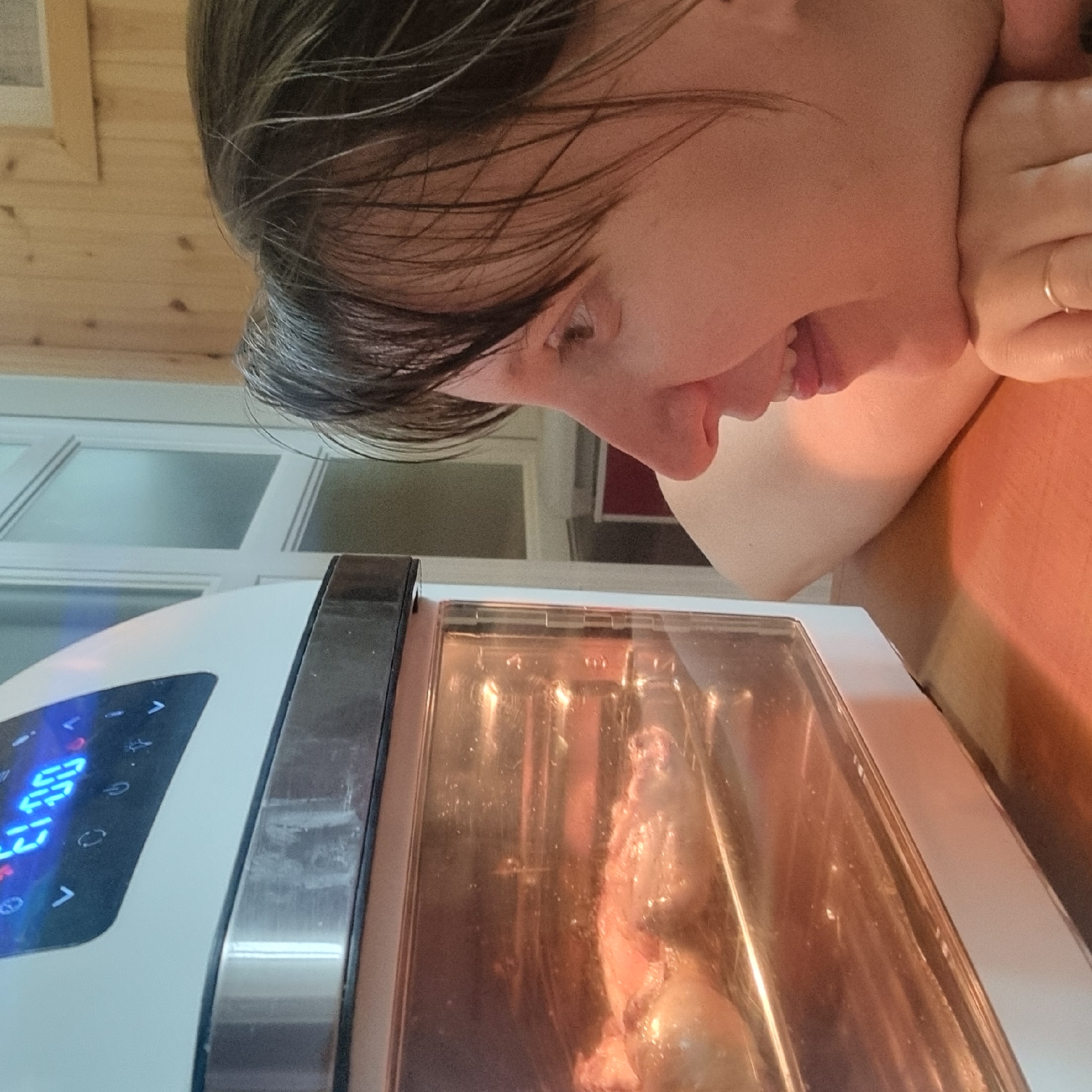 An adult, identified by their skin tone and facial features, is positioned in the foreground, looking at an oven. The oven, a white and metallic appliance, contains food, likely chicken, being cooked. The oven's digital display shows "21:00". The background features a wooden paneled wall and a glass door with horizontal dividers, suggesting an indoor domestic setting. The overall lighting is warm and illuminated by the oven's interior light. The image is captured from a medium close-up perspective, focusing on the adult's face and the oven. There are no visible indications of the city of Dunnae-myeon, South Korea.