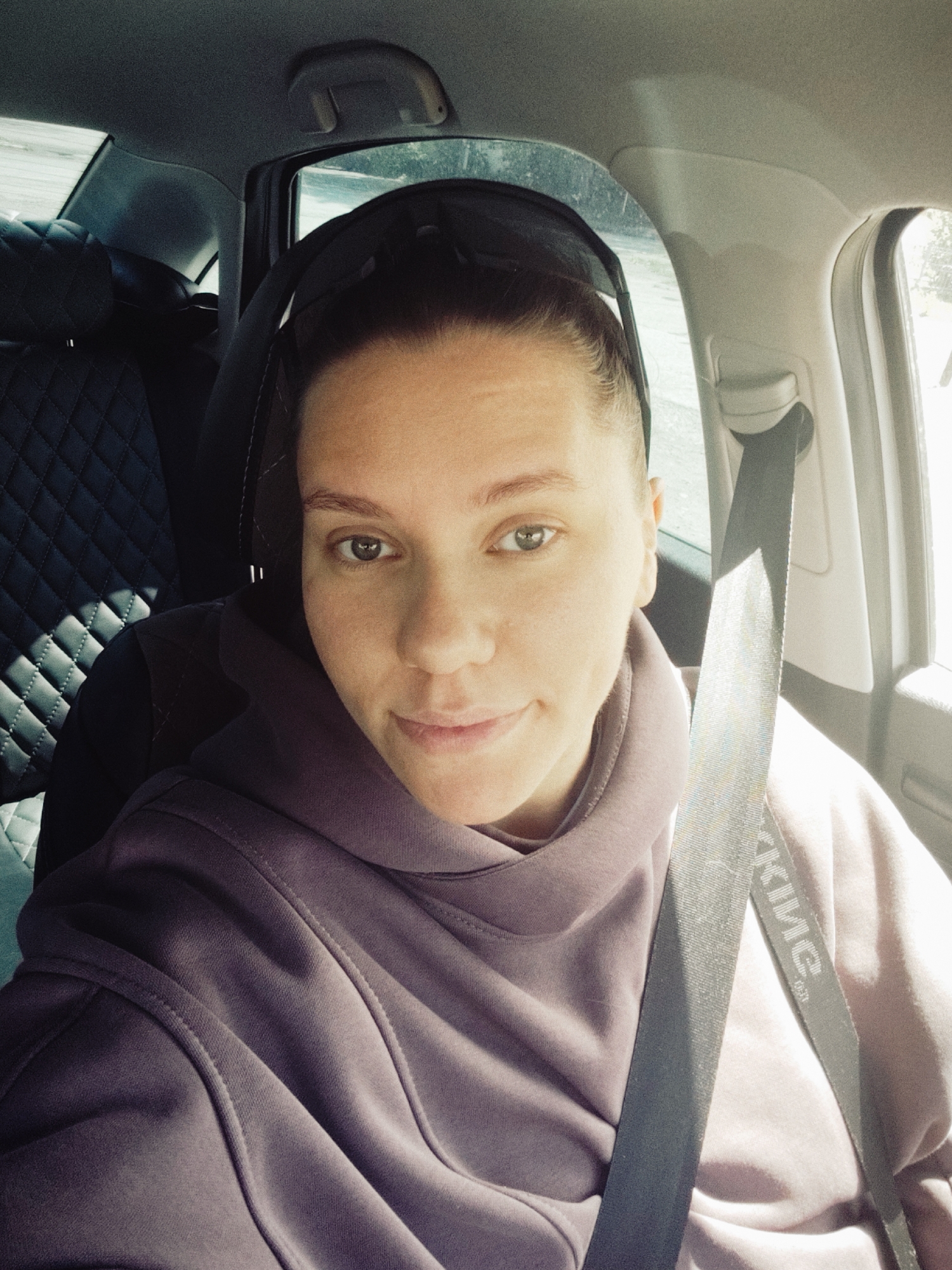 A woman is seated in the driver's seat of a car, wearing a purple hooded sweatshirt and a seatbelt. The car has dark leather seats with a diamond-stitched pattern. The interior is visible, including the headrest and the car's ceiling. The background shows a glimpse of the outside environment through the car window, possibly a road and foliage. The lettering visible on the seatbelt reads "ОКИСЕ". The woman is looking directly at the camera. The location is Chaikovsky, Russia.