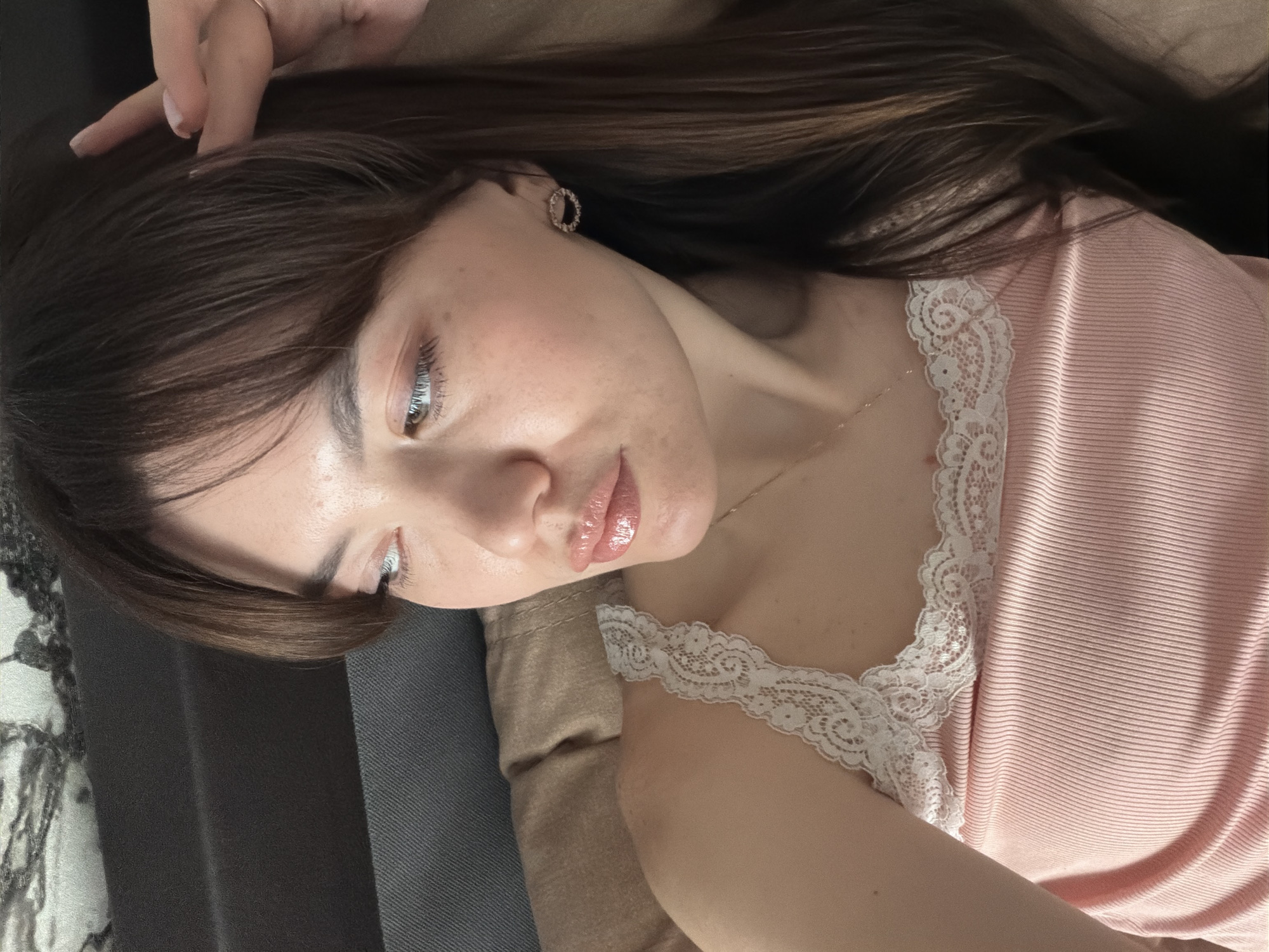 A young woman with brown hair and light skin is lying down. She is wearing a pink top with white lace trim around the neckline. She has earrings in her ears. Her face is angled towards the camera, and she is looking down. Her lips appear glossy. She is lying on what appears to be a sofa with different colored sections. The location is not identifiable as Chaikovsky, Russia from the visual information provided.