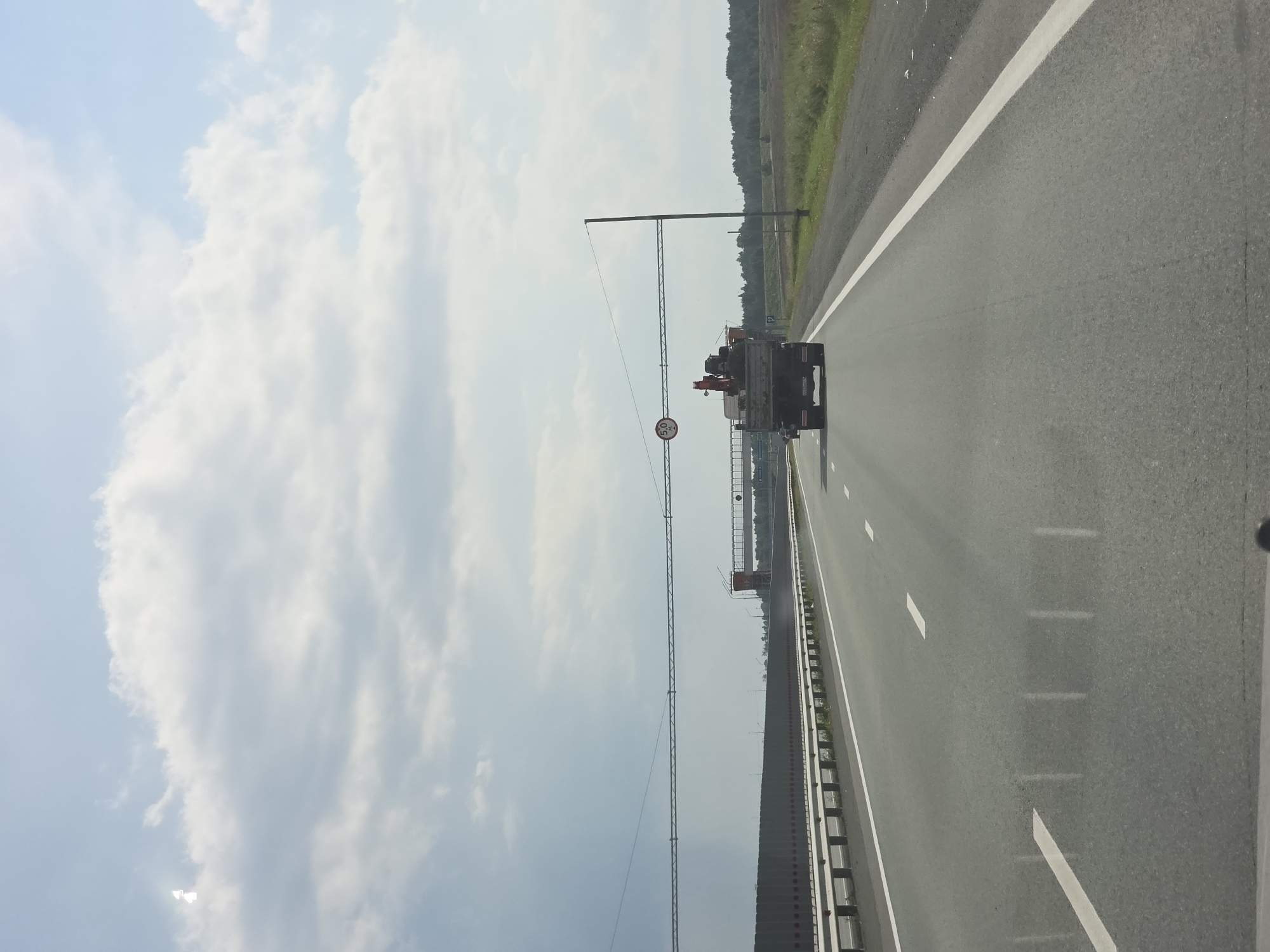 The image depicts a highway scene, likely in or near Borisovka, Russia, under a partly cloudy sky. In the foreground, the asphalt road is visible with white lane markings. A guardrail runs along the right side of the road. In the middle ground, a truck with a crane on its bed is traveling away from the viewer. Above the road, a gantry supports a circular sign indicating a height restriction of 5.0 meters. Further in the background, a sound barrier runs along the left side of the highway, and a line of trees marks the horizon. The overall environment suggests a rural or semi-rural highway setting.