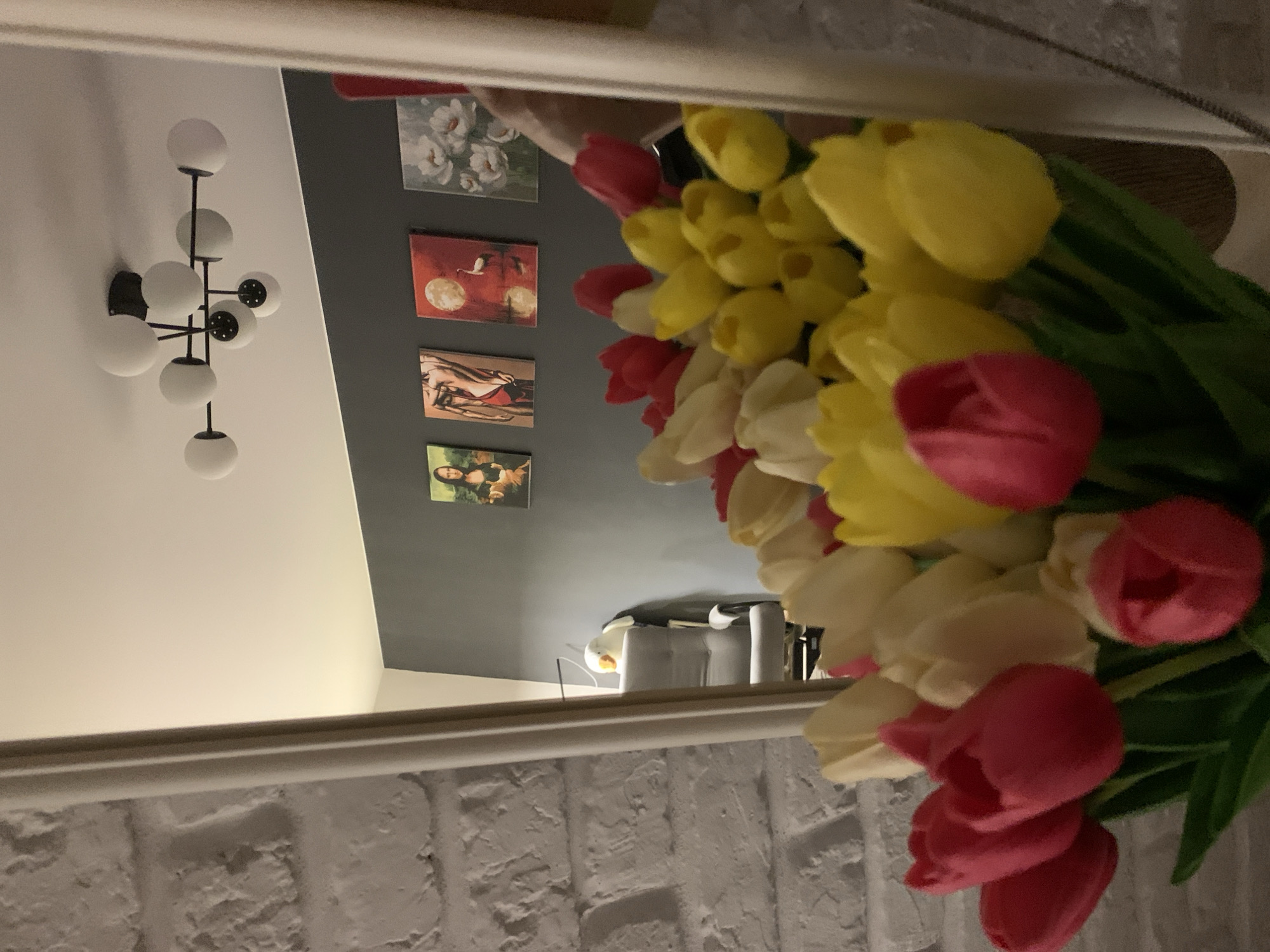 A bouquet of artificial tulips, predominantly red, yellow, and white, is reflected in a mirror. The tulips are arranged in the foreground and fill the lower half of the image. Behind the tulips, the reflection shows a grey wall adorned with four framed pictures. From left to right, the pictures depict: a portrait of Mona Lisa, a figure of a woman, a scene with a crane and a moon over water, and a cluster of white flowers. Above the pictures, a modern black chandelier with multiple white spherical lights is visible on the ceiling. To the left of the pictures, a light grey armchair is partially visible. The mirror itself has a light-colored frame. The wall on the left side of the image, outside the reflection, is a white brick wall. The scene is likely an indoor residential setting. There are no people present in the image. The city of Kapyĺ, Belarus, is not indicated in the image.