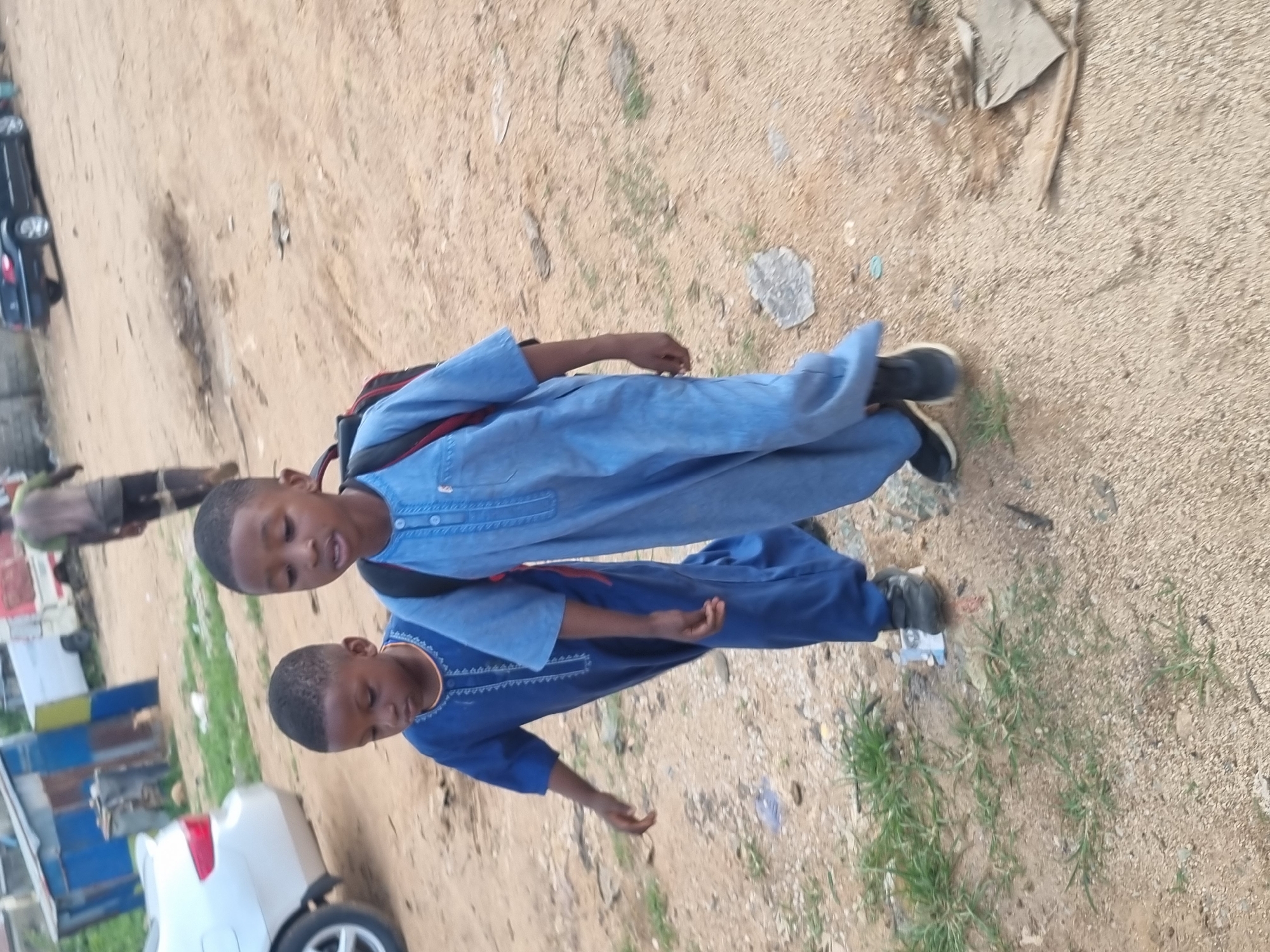 In the city of Jos, Nigeria, two young boys, likely brothers, walk side-by-side on a dirt road. The boy in front, wearing a blue robe and a backpack, has a serious expression as he walks. Behind him, his younger brother, also in a blue robe, looks down at his feet. The road is dusty and uneven, with patches of dry grass and scattered debris. In the background, a white van and a silver car are parked, suggesting a residential or commercial area. The overall scene depicts a typical day in Jos, with children playing and going about their daily routines.