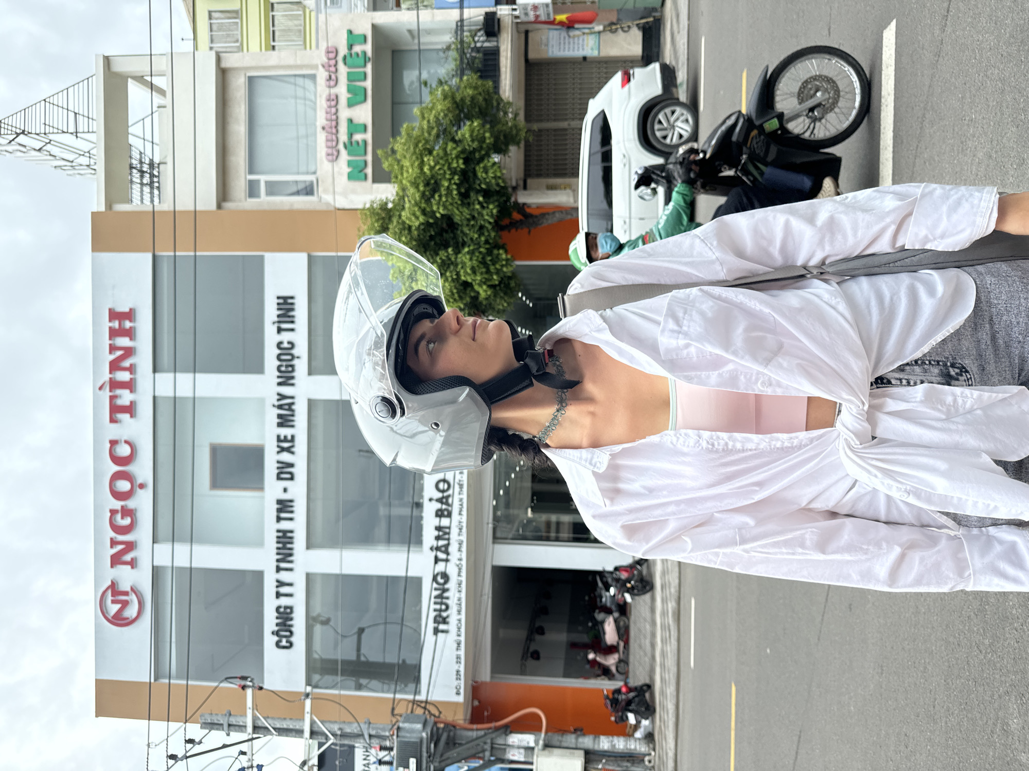 Here's a description of the image:
A woman wearing a white helmet and a partially unbuttoned white shirt over a light pink top is standing on a city street. The background features a large building with "NT NGỌC TỈNH" vertically displayed in red letters on a white section. Below this, in smaller black letters, is "CÔNG TY TNHH TM - DV XE MÁY NGỌC TỈNH". Further down the building is "TRUNG TÂM BẢO" with an address "ĐC: 229 - 231 THỦ KHOA HUÂN KHU PHỐ 1 PHỦ THỦY - PHAN THIẾT". Another sign in green text reads "NÉT VIỆT." A white car and a scooter are visible on the street behind her. The photo appears to be taken in Phan Thiết, Vietnam, based on the address.
