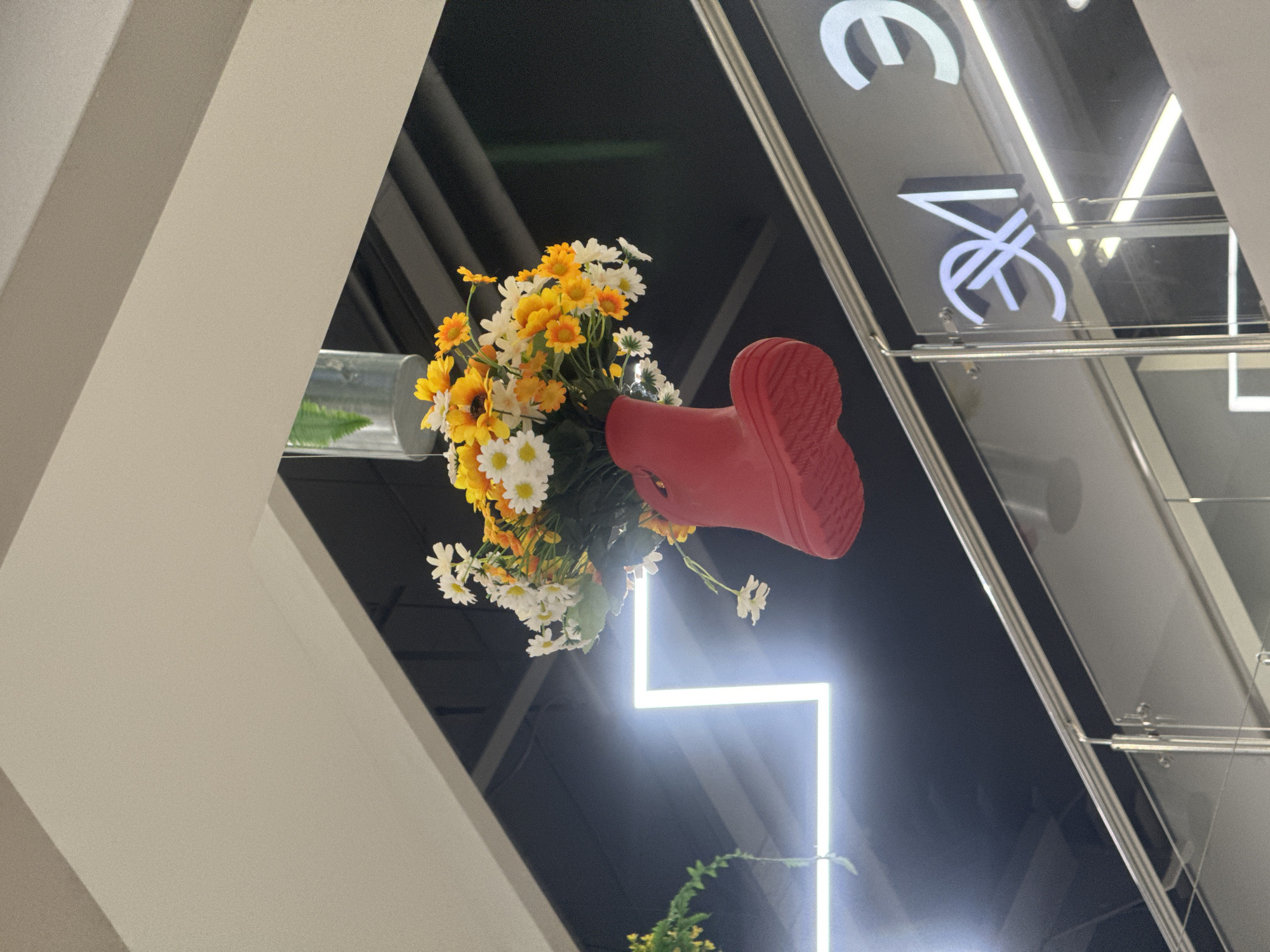 A red Wellington boot filled with a bouquet of white and yellow artificial daisies hangs suspended from the ceiling indoors, with a clear plastic container visible inside the boot holding the flowers. Below the boot, a reflective railing separates different levels of the interior. A large, illuminated white square light fixture is mounted on a dark ceiling in the background. To the right, a business logo is visible on a glass surface, featuring white stylized lettering on a dark background. The environment appears to be a commercial space, possibly a shopping mall or retail store. The exact location in Irkutsk, Russia, cannot be determined from the visual information provided.