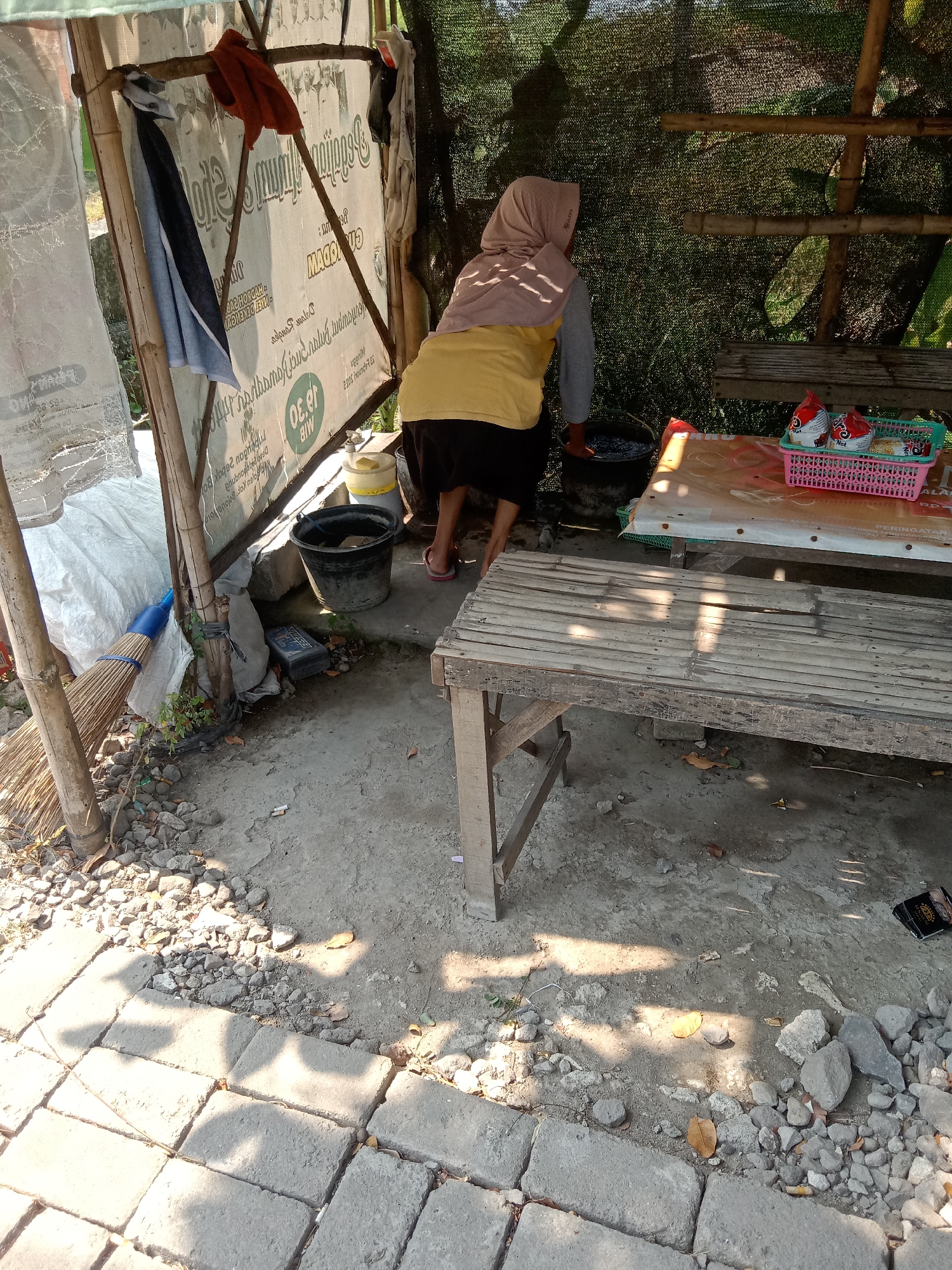In Kemamang, Indonesia, a woman wearing a light brown hijab and a yellow shirt over dark pants is seen from behind. She is bending over, with her right hand in a black bucket filled with water. To her left is a broom with a blue handle leaning against a bamboo structure covered with a white tarp. The ground is a mix of concrete pavers and dirt, scattered with rocks and dried leaves. A bamboo bench is positioned in the foreground, and a table covered with a patterned cloth is visible behind the bench. On the table, a pink basket holds several red and white packaged items, likely food products. The scene appears to be an outdoor area, possibly a roadside stall or a small outdoor workspace, with a partially visible bamboo structure and netting in the background, suggesting a covered or partially enclosed space. Sunlight casts shadows on the ground, indicating it is daytime.