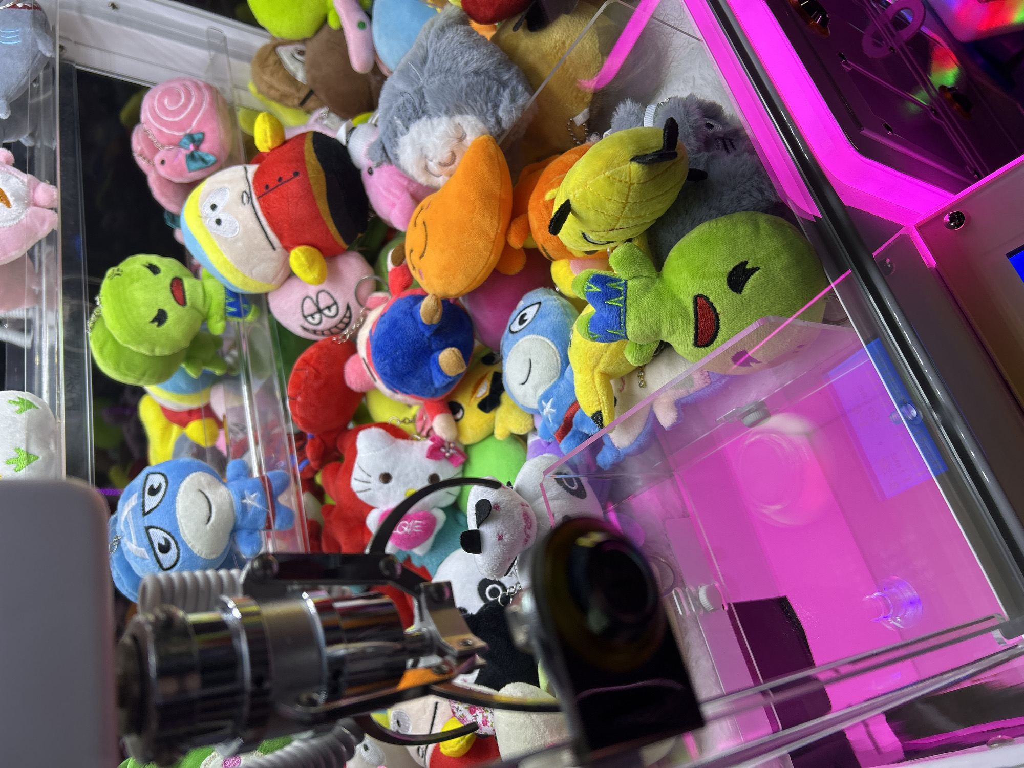 The image provides a close-up view inside a brightly lit claw machine, filled to the brim with numerous colorful plush toys. The scene is set in an entertainment venue. The machine's transparent plastic walls reflect a vivid pink and purple LED light from within, casting a glow over the prizes.
In the foreground, a metallic claw mechanism is visible, angled towards the lower-left, partially obscuring some of the toys below. The plushies are densely packed, ranging from recognizable cartoon characters to generic cute animals and shapes.
Notable characters include an Eric Cartman plush from South Park, distinguished by his red, yellow, and blue outfit, positioned towards the center. A white Hello Kitty plush, adorned with a pink bow, holds a red heart with the word "LOVE" printed on it. A green alien plush, reminiscent of the "Little Green Men" from Toy Story, with blue accents on its hands, is also clearly visible. Another green plush with a wide, open red mouth is situated more towards the right. Other diverse plushies include a pink snail-like creature with a blue bow, a grey fluffy animal, an orange teardrop shape, and a blue creature with large, round eyes. A black and white panda plush and a small white plush keychain with the text "GIE" are also discernible among the collection.
The image captures the vibrant and exciting atmosphere typical of an arcade game.