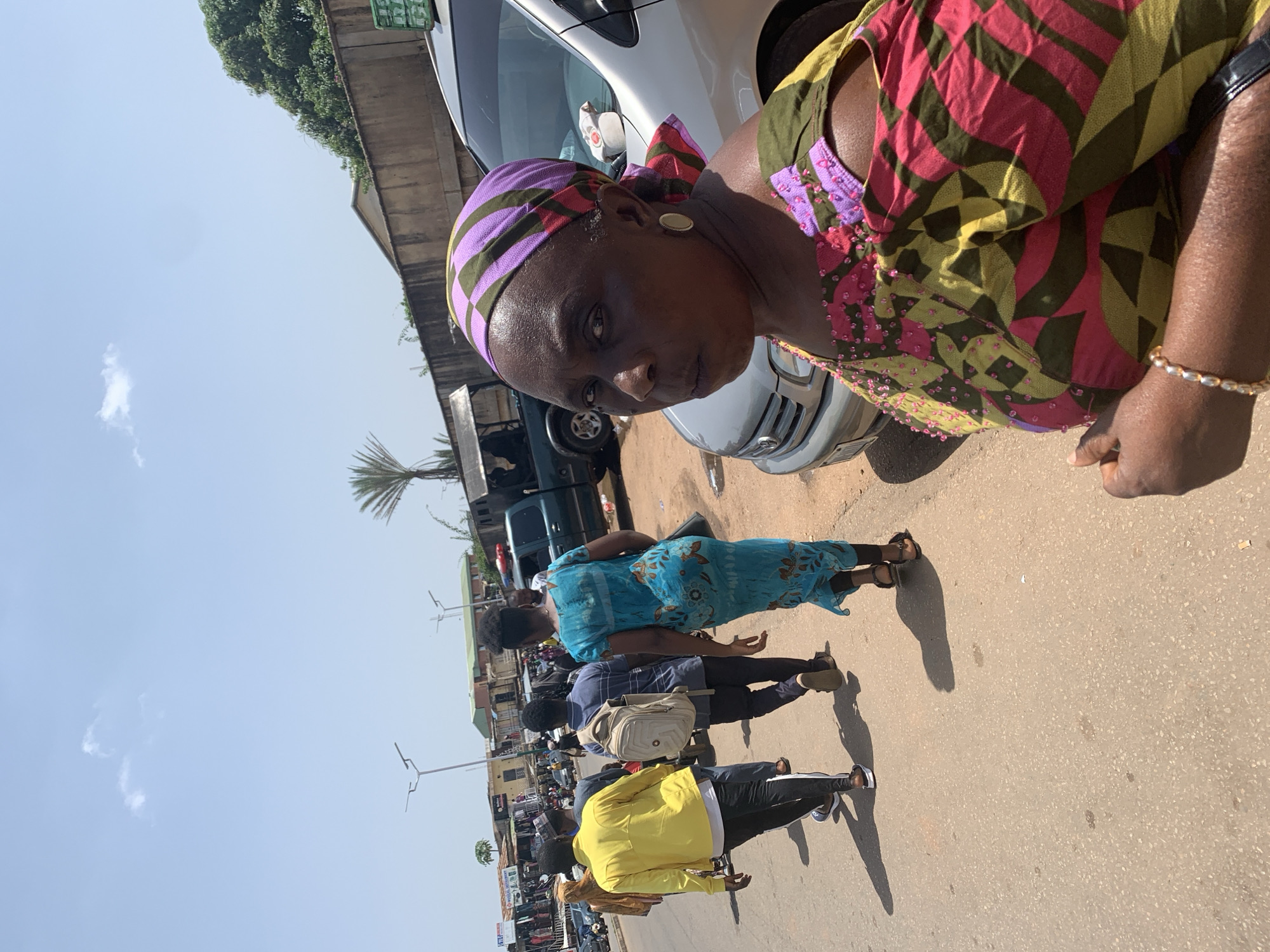 The image shows a woman wearing a colorful headscarf and a patterned dress, looking directly at the camera. She is positioned in the foreground, with a silver vehicle and other people in the background. The scene appears to be an outdoor market or street in Jos, Nigeria, indicated by the architecture and vehicles. Several individuals are walking in the street, some carrying bags or backpacks. The weather seems sunny with a clear sky. The overall impression is that of a busy, everyday scene in a Nigerian urban environment.