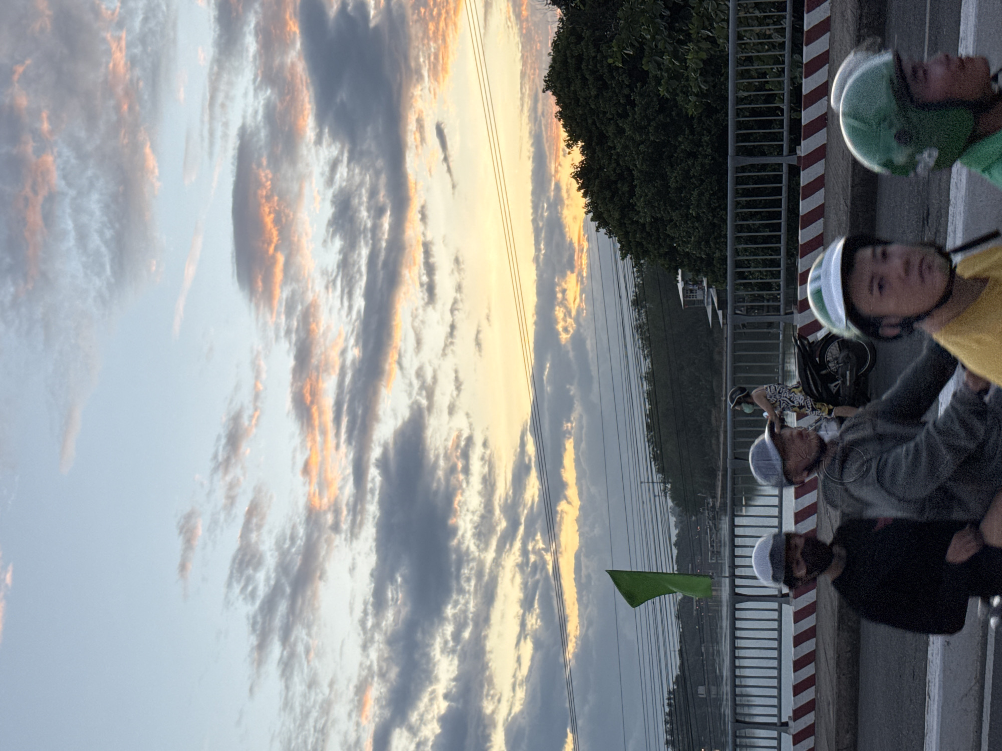 This image captures a sunset scene on a bridge in Nha Trang, Vietnam, likely near the Xóm Cồn area. The sky is a canvas of soft blues and grays, streaked with vibrant orange and pink hues from the setting sun. Wispy clouds create a textured overlay on the horizon. A lush green tree line forms a dense backdrop to the bridge, with a body of water visible beyond the railing.
On the bridge, several individuals are riding on motorcycles. In the foreground, a person wearing a black shirt and a face mask is seated behind the rider of another motorcycle. The rider in front is wearing a gray jacket and a helmet. To the right, two individuals are on a motorcycle, both wearing helmets. One is wearing a yellow shirt and the other a green helmet. Further back, a person in a patterned top is also on a motorcycle. A green flag is visible on a pole on the left side of the bridge. Power lines crisscross the upper portion of the image, adding a functional element to the natural beauty of the sunset. The overall atmosphere suggests a typical evening commute on a bridge in Nha Trang.
