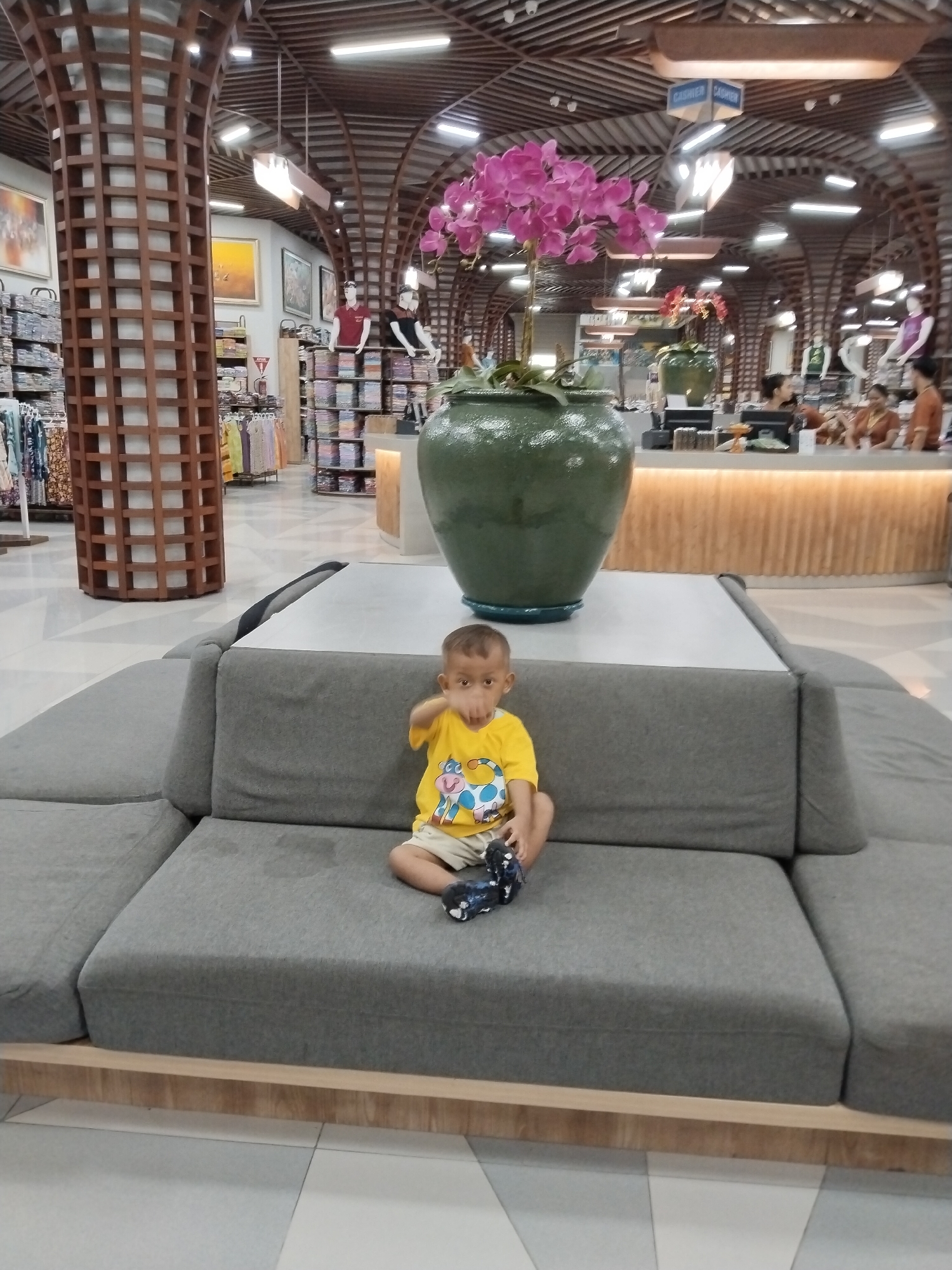 A young boy wearing a yellow t-shirt and light-colored shorts sits on a grey sectional sofa in what appears to be a retail store. The boy is looking towards the camera with his right hand extended. He is holding a dark-colored toy in his left hand. In the background, shelves filled with clothing and display mannequins are visible. A large green planter with a pink orchid is positioned behind the sofa. The store's interior features wooden architectural elements, including curved pillars and ceiling structures. A counter with cashiers is also visible in the background. The floor is tiled. This scene is likely in Kuta, Indonesia, given the presence of a retail store typical of tourist areas there.