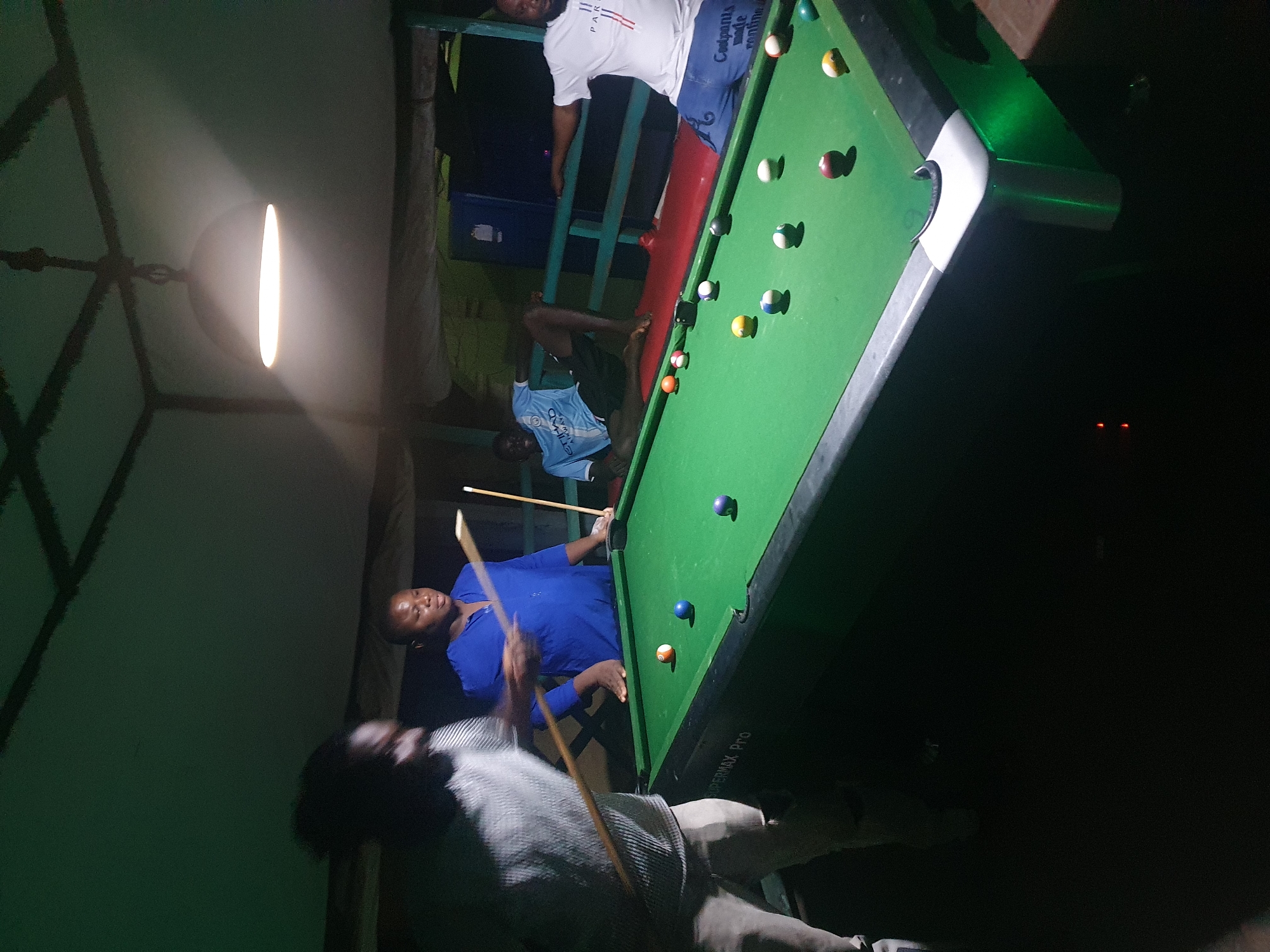 The image depicts a group of four individuals engaged in a game of billiards in an outdoor, possibly covered, setting. The scene appears to be a local establishment in Lokoja, Nigeria, suggested by the ambiance and the presence of a billiard table and players.
In the foreground, a person is blurred due to motion, holding a billiard cue and positioned near the billiard table. To their right, a person wearing a blue shirt is actively participating in the game, holding a cue and looking towards the table. In the background, two more individuals are visible. One, wearing a light blue and white football jersey, is seated and appears to be observing the game. The other, wearing a white t-shirt and blue shorts, is also seated and facing towards the table.
The billiard table is green with various colored balls scattered across its surface. The environment is dimly lit, with a hanging light fixture providing illumination, casting shadows and creating a somewhat atmospheric feel. The overall scene suggests a casual and social gathering centered around the game of billiards.