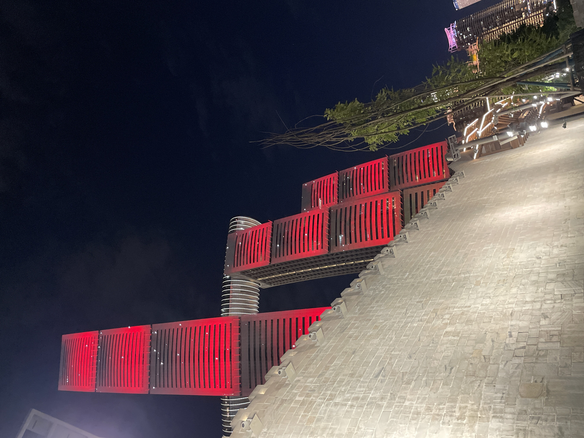 An eye-level, wide-angle, low-light photograph captures the Hongyadong complex in Yuzhong District, China, at night. The iconic architecture features multiple towers illuminated in vibrant red, with horizontal bracketing visible on each level. The tallest tower stands to the left, appearing to be connected to a neighboring tower by a curved, white-lit structure. To the right, another tower, also red-lit, is partially visible. In the foreground, a textured, light-colored stone wall dominates the lower portion of the frame, with small, dark lights mounted along its top edge. Behind the wall, a cluster of slender, bare tree branches extends upwards. Further in the background, glimpses of other city buildings are visible, some also lit at night, contributing to the urban landscape. The sky is dark blue, with faint clouds. No people are visible in the scene.