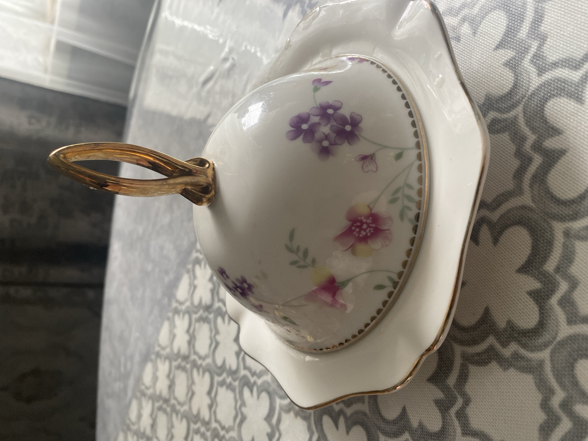 A lidded dish with floral patterns and a gold handle sits on a patterned tablecloth. The dish is white with pink and purple flowers. The tablecloth has a gray and white geometric pattern. The background is blurred and appears to be indoors. There is no indication of the location being Chontaul, Russia, or any other city.
