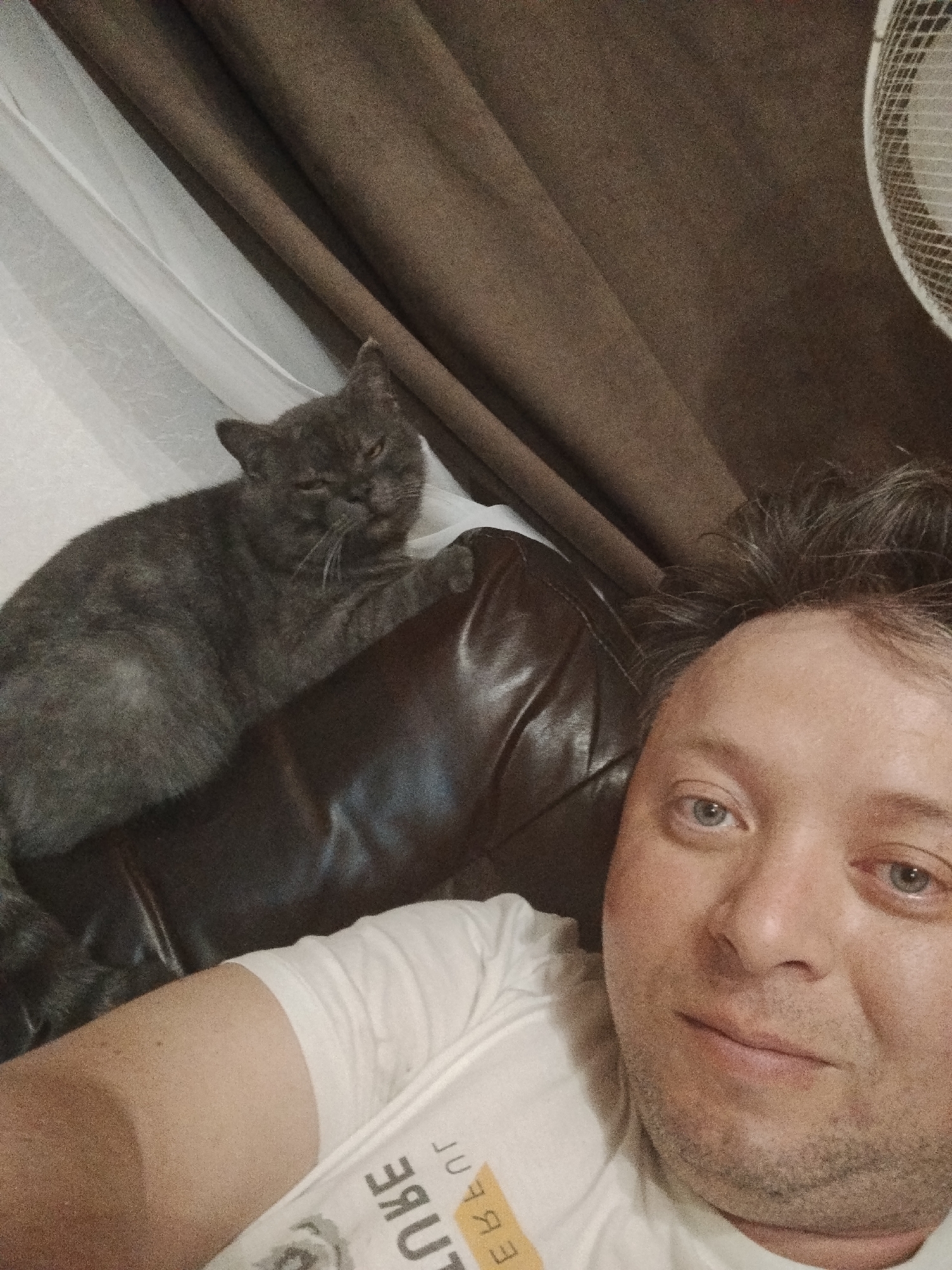 A selfie taken from a low angle shows a man with blue eyes and unkempt hair lying on what appears to be a leather couch. He is wearing a white t-shirt. Resting on the couch to the left is a gray cat with its eyes partially closed. Behind the cat and the man are curtains, with a section appearing to be a light color and another part a darker brown. A partial view of a fan is visible in the upper right corner. The location is Unknown, Unknown.