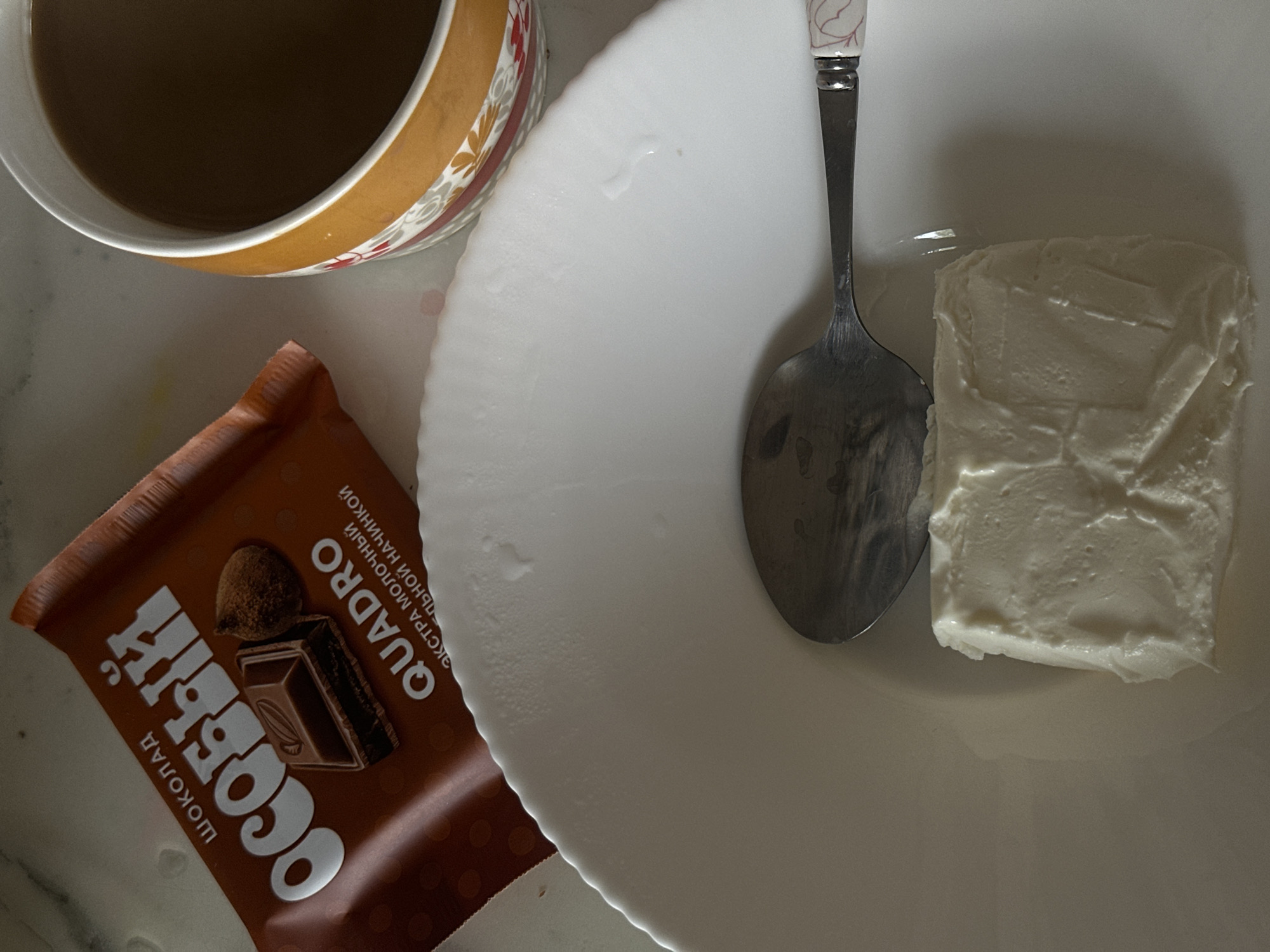 The image shows a flat-lay of a snack and drink arrangement. There is a brown chocolate bar labeled "ОСОБЫЙ" and "QUADRO" in Russian. Next to it is a white plate with a metal spoon and a block of white cream cheese. A cup with a brown liquid, likely coffee or tea, is positioned at the top left corner. The scene appears to be set on a light-colored countertop. Location: Черёмушки, Russia is speculative and cannot be confirmed from the image alone.