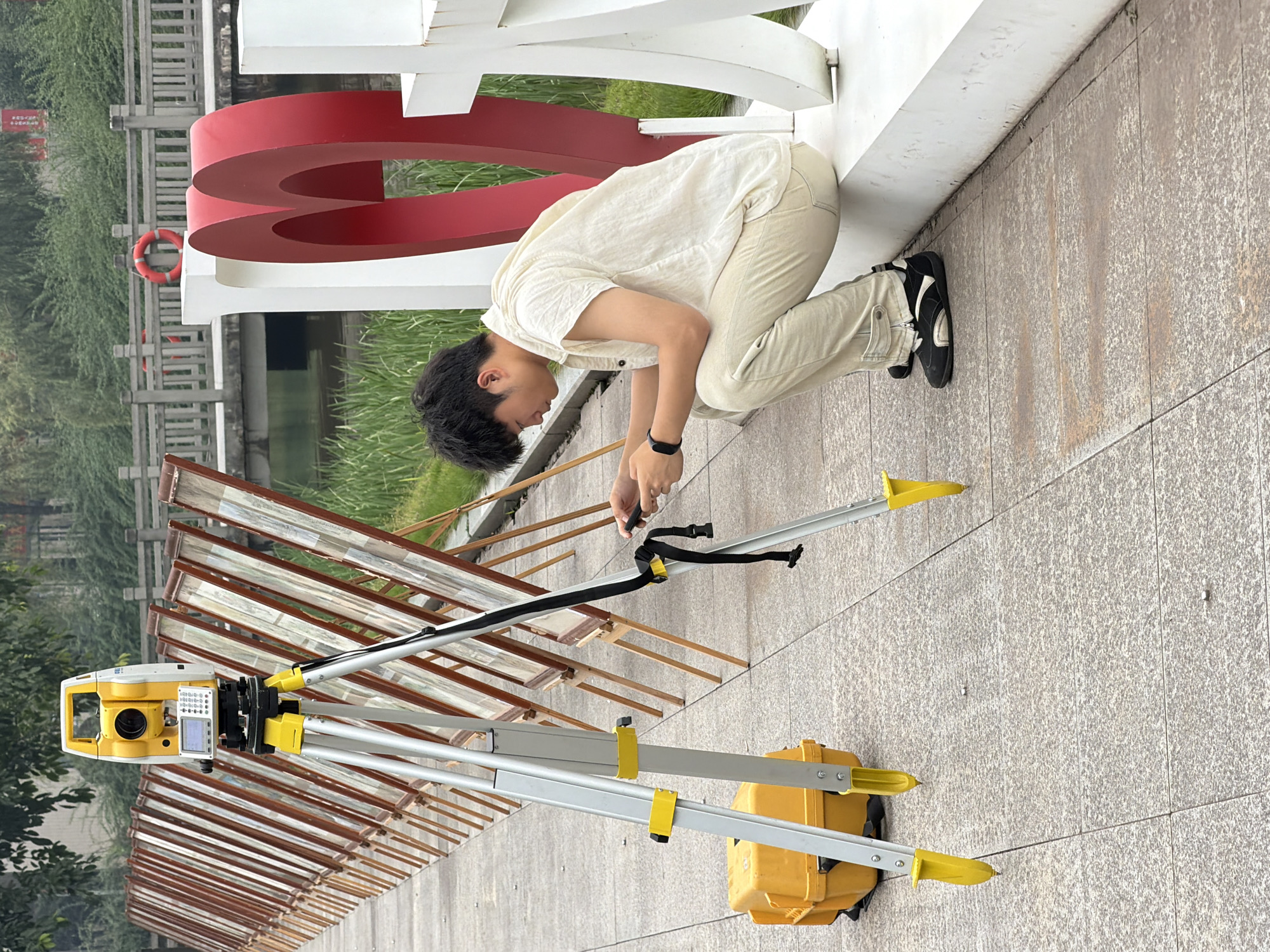 A young man is squatting on a white ledge in front of a large red heart sculpture, looking at his phone. He is wearing a light-colored short-sleeved shirt and light-colored pants with cargo pockets. Next to him, a surveying instrument is set up on a tripod, with a yellow carrying case on the ground nearby. The surveying instrument is a yellow and white total station, commonly used in construction and land surveying. In the background, a row of framed artwork is displayed on easels along a pathway next to a body of water. A wooden bridge with railings spans the water in the distance. The scene appears to be outdoors, possibly in a park or public area, in the city of Jiangjin, China.