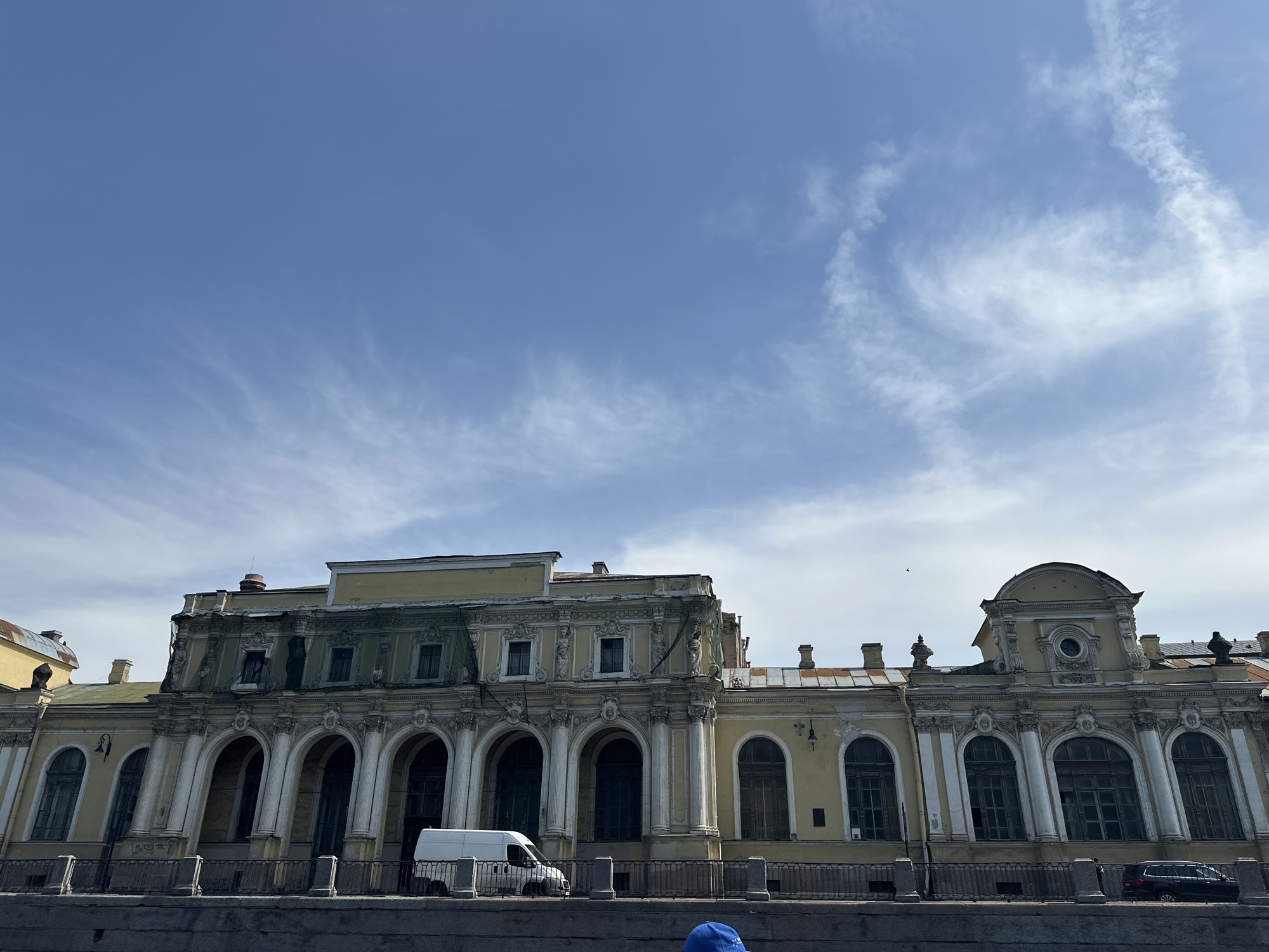 The image shows a large, yellow, ornate building in Saint Petersburg, Russia. The building appears to be in need of repair, as evidenced by netting and discoloration on the façade. The architecture features numerous arched windows and decorative elements such as columns and sculptures. A white van is parked in front of the building, near what seems to be a canal or river. Part of a blue object is visible at the bottom center of the frame. The sky is blue with wispy clouds. A black car is parked beside the building and a bird is flying overhead. The image is taken from a slightly low angle.
