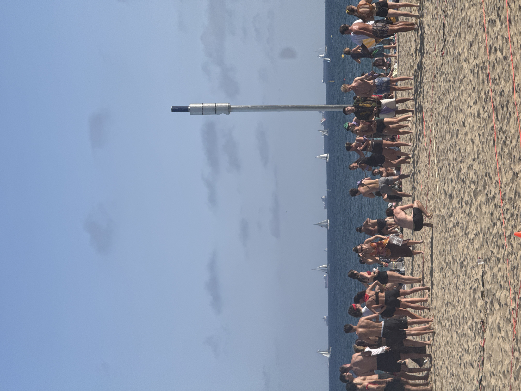 This is an image of a crowded beach in Barcelona, Spain. The sky is blue with some clouds, and the sea is visible in the background. Numerous sailboats and cargo ships are scattered across the water. On the beach, a large group of people are gathered, many in swimwear, suggesting a warm day. Some individuals appear to be interacting, while others are standing or sitting. A tall, metallic pole with lights is a prominent feature on the beach. Red and white ropes are laid out on the sand, possibly indicating a boundary or a game. The sand is light brown and appears to be a typical beach environment.