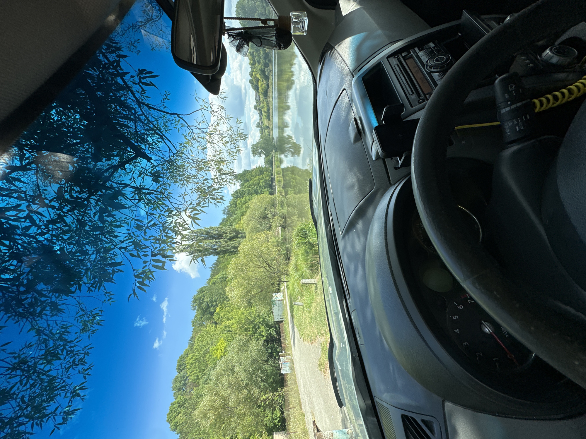 The image depicts the view from the driver's seat of a car parked near a body of water. The car's dashboard and steering wheel are visible in the foreground. Through the windshield, a serene natural landscape unfolds. Lush green trees line the banks of a calm lake, which reflects the blue sky and scattered clouds. A dirt road leads away from the car towards the left, bordered by grassy terrain. In the distance, there are hints of structures and more rolling hills covered in foliage. The scene suggests a peaceful outdoor setting, possibly a recreational area on the outskirts of Makiv, Ukraine, given the context provided. There are no people visible in the image.