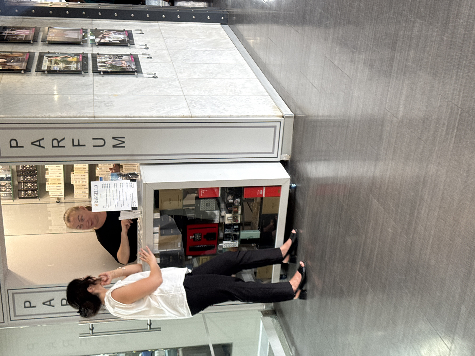 Two women are standing at a counter in a retail store. The woman on the left, wearing a white sleeveless top and black pants, is looking at the woman on the right. The woman on the right, wearing a black top, is facing the woman on the left and appears to be speaking to her while looking at some documents on the counter. Behind them, shelves are stocked with various perfume bottles and boxes. The store has a marble-tiled wall on the right with framed pictures displayed. The floor is a polished grey tile. The store appears to be a perfume shop located in Yalta, Ukraine, with a sign on the left pillar reading "PAR" vertically.
