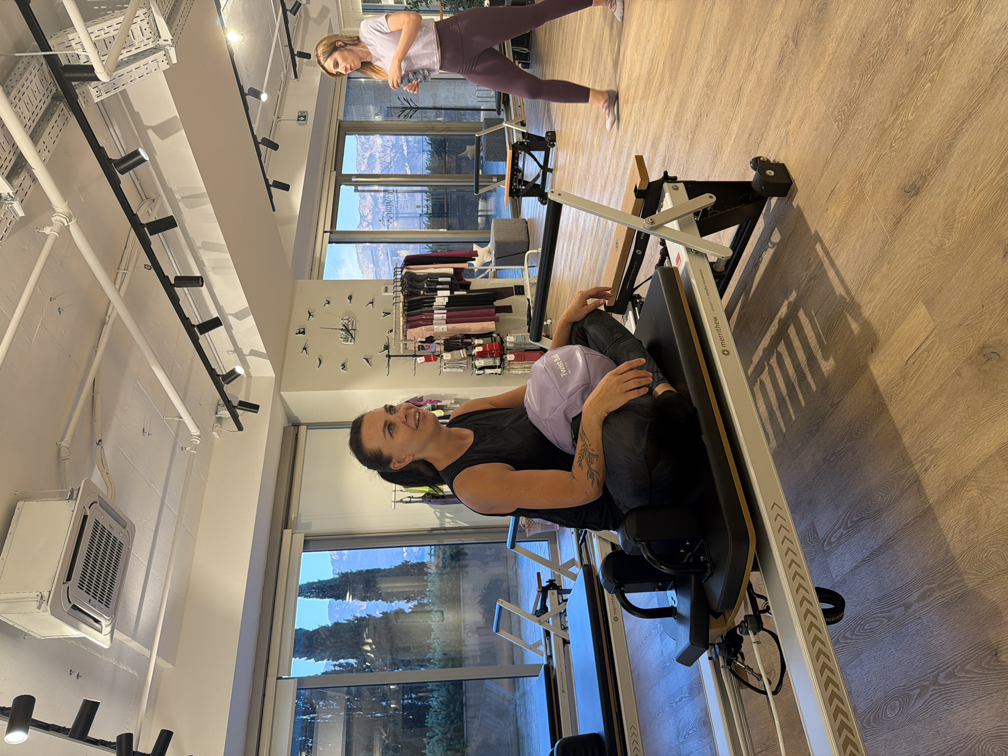 In a well-lit fitness studio located in Boreti, Montenegro, two women are engaged in activity. The foreground features a woman sitting on a Pilates reformer, holding a light purple ball. She is wearing a black tank top and grey patterned leggings. Her left arm bears a tattoo of a branch with leaves. The Pilates reformer is a Merrithew brand, with metal and wood components. Behind her, another woman stands, holding a clear water bottle. She is wearing a light purple crop top and dark purple leggings. She is facing away from the camera and appears to be walking. The studio has large windows that look out onto a landscape of trees and mountains. The room has a wooden floor and a suspended ceiling with track lighting and an air conditioning unit. In the background, clothing racks display activewear, and other fitness equipment, including additional Pilates reformers and chairs, is visible.