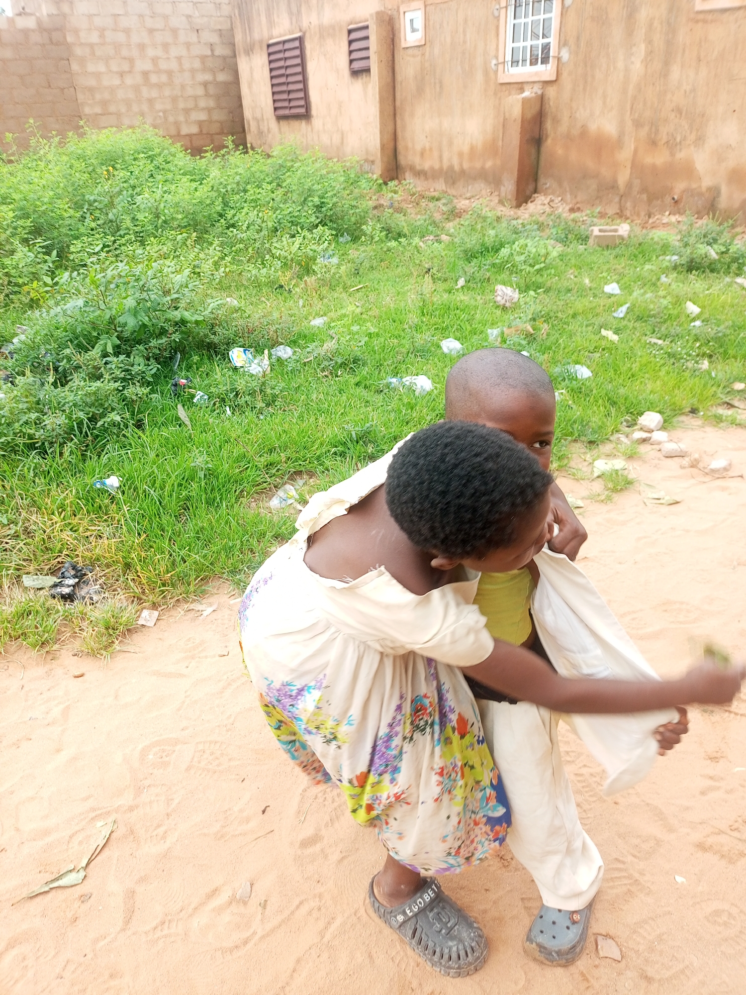 In Tempure, Nigeria, two young children are depicted outdoors. The child in the foreground, facing away from the camera, is being carried on the back of another child. The child being carried is wearing a light-colored dress with a floral pattern. The child carrying has on light-colored pants. Both children are barefoot. The ground is sandy, with patches of green grass and some scattered debris. In the background, a building with a brick facade and windows can be seen. The overall impression is of a rural or semi-urban setting in Nigeria.