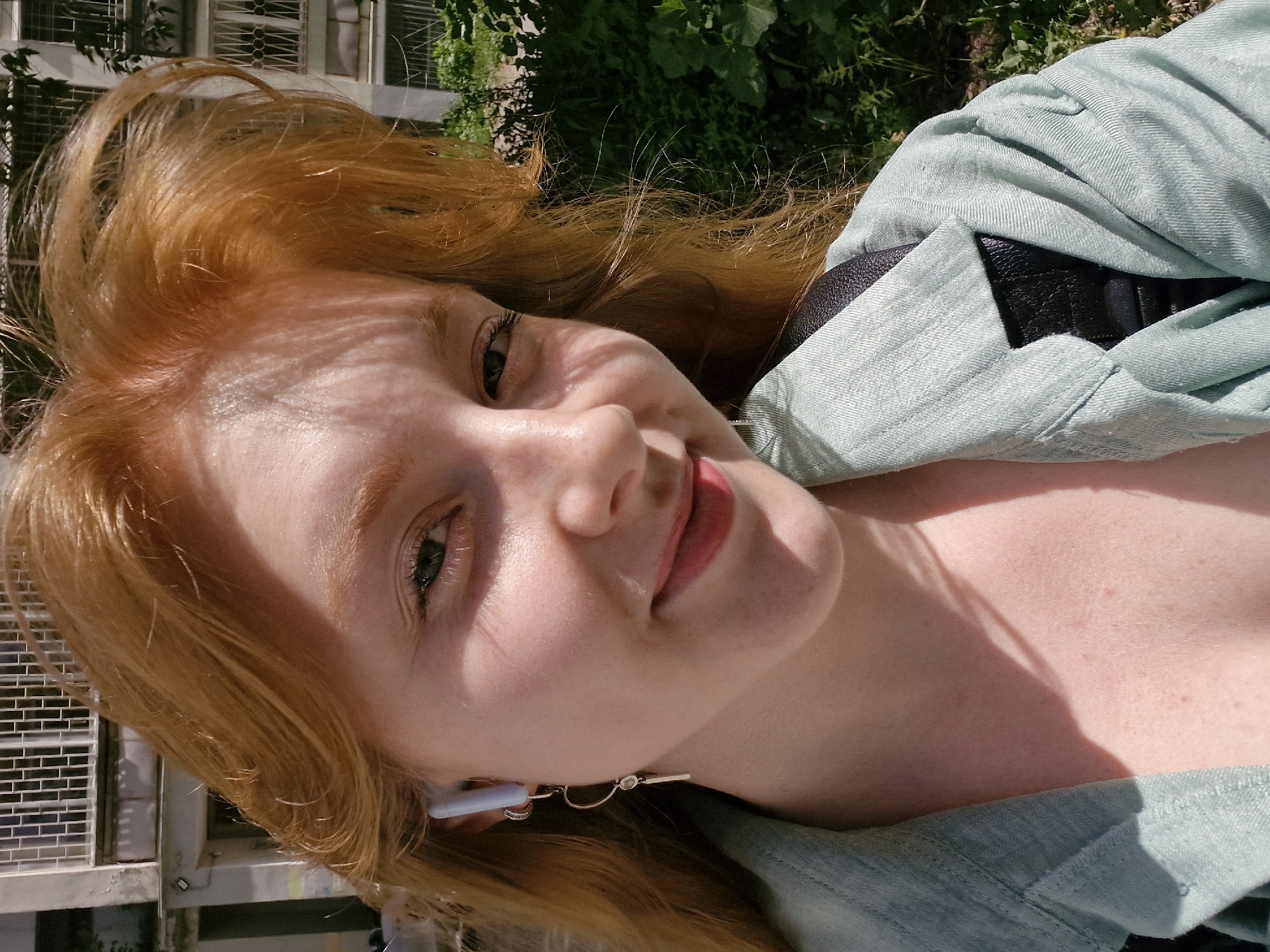 A young woman with red hair is outdoors, possibly in the city of Yekaterinburg, Russia, based on visual cues such as building style. She is wearing light blue clothing and a black backpack. She has an AirPod in her left ear. She is likely near trees and a building, which cast shadows on her face.