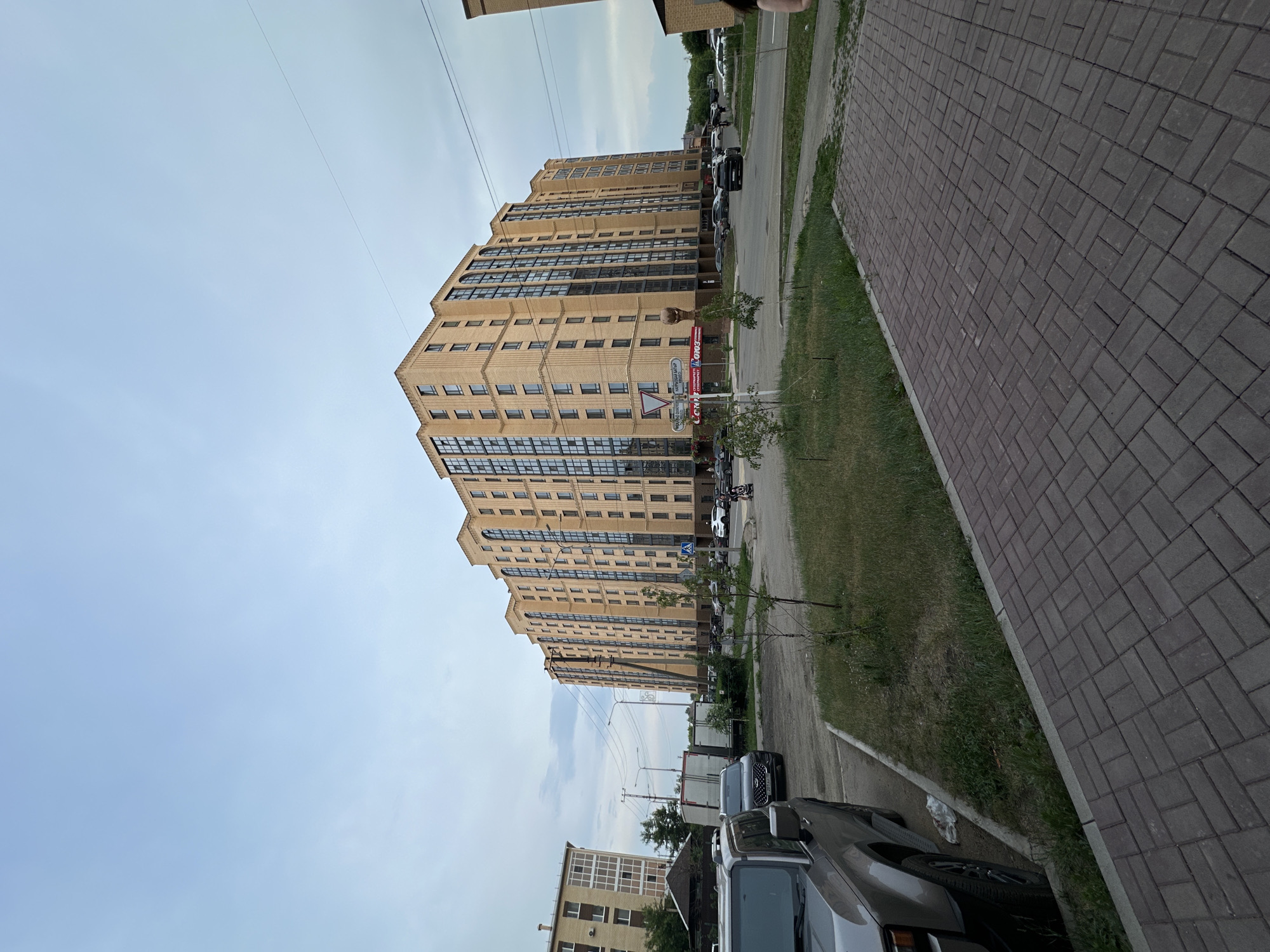 The image depicts a multi-story residential building in Kokshetau, Kazakhstan, under an overcast sky. The building, with its yellow-beige brick facade and numerous dark-paned windows, dominates the center and right side of the frame. In the foreground, a gray SUV is parked on the left. A paved walkway with a brick pattern runs across the bottom of the image. Beyond the SUV, there are other vehicles and a smaller, lighter-colored building. The area appears to be a residential or mixed-use urban environment with some greenery and street furniture, including traffic signs.